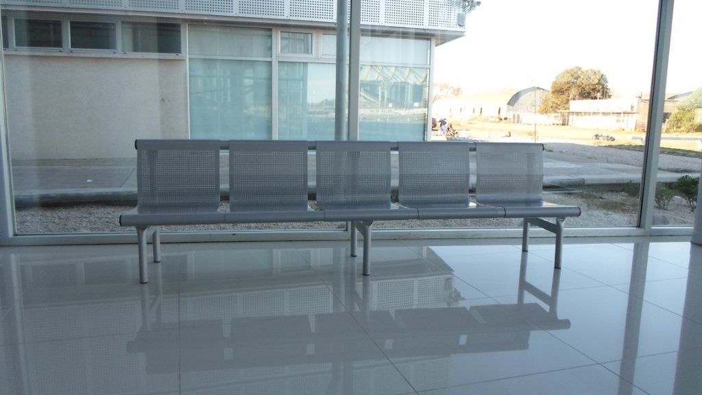 Banco de metal gris en un luminoso vestíbulo interior con suelo de baldosas brillantes y paredes de cristal.