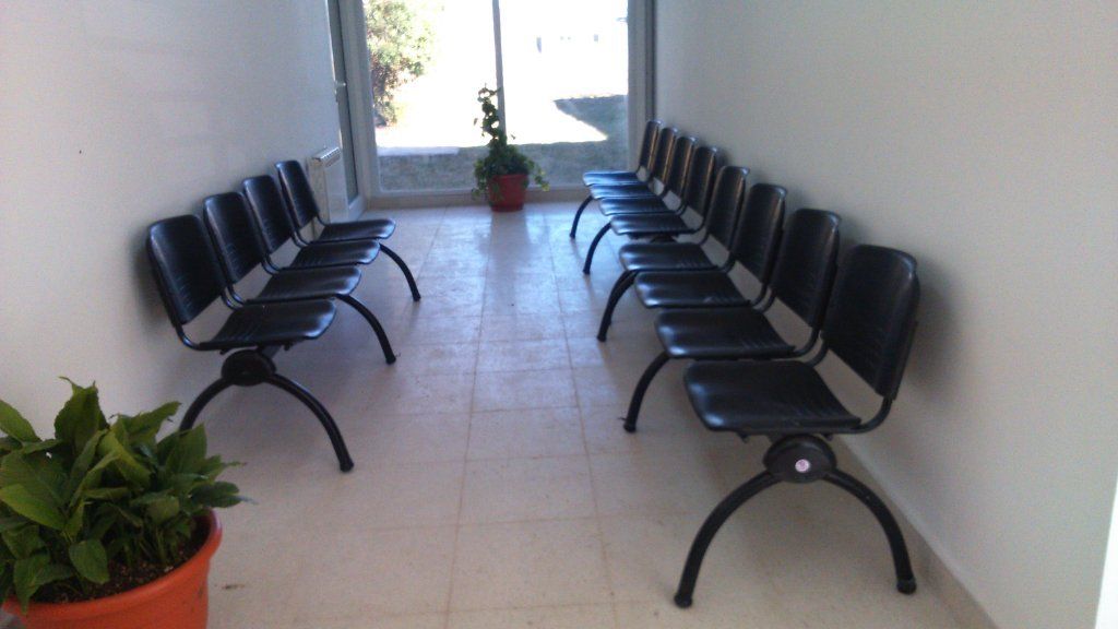 Zona de espera con filas de sillas negras frente a una ventana, con plantas en macetas en ambos extremos.