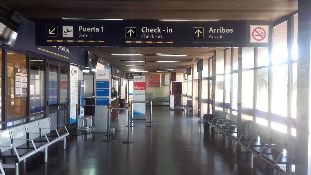 Pasillo vacío de la terminal del aeropuerto con letreros de facturación y llegadas, filas de asientos y ventanas luminosas.