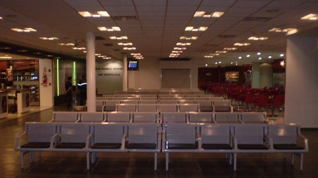 Sala de espera vacía del aeropuerto con filas de asientos grises bajo luces brillantes del techo.