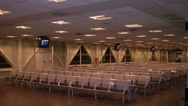 Sala de espera vacía del aeropuerto con filas de asientos y monitores en el techo.