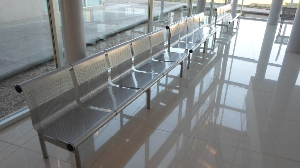 Fila de bancos metálicos de espera del aeropuerto junto a grandes ventanales en un luminoso pasillo de la terminal.