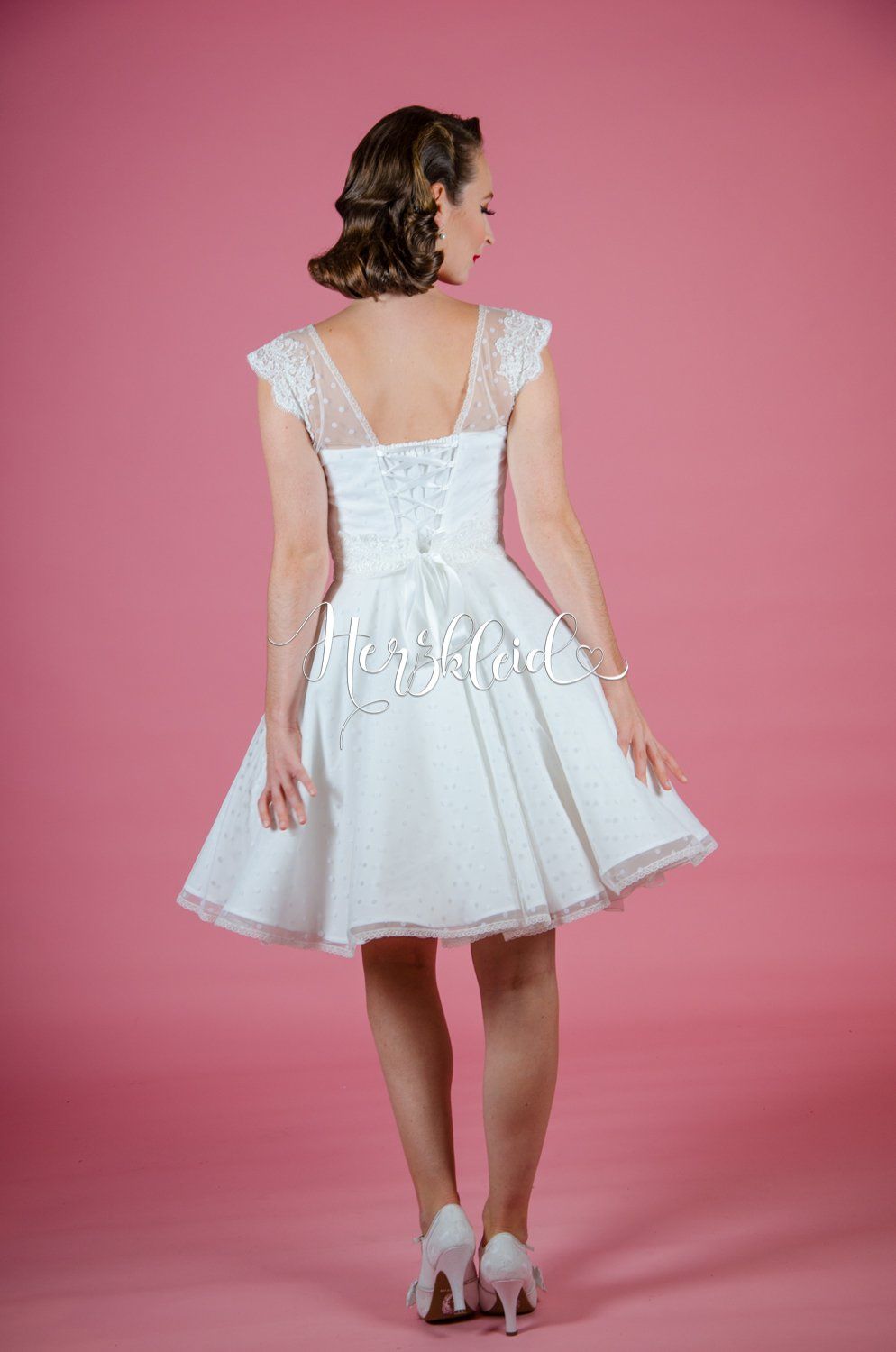 Eine Frau in einem weißen Kleid steht vor einem rosa Hintergrund.