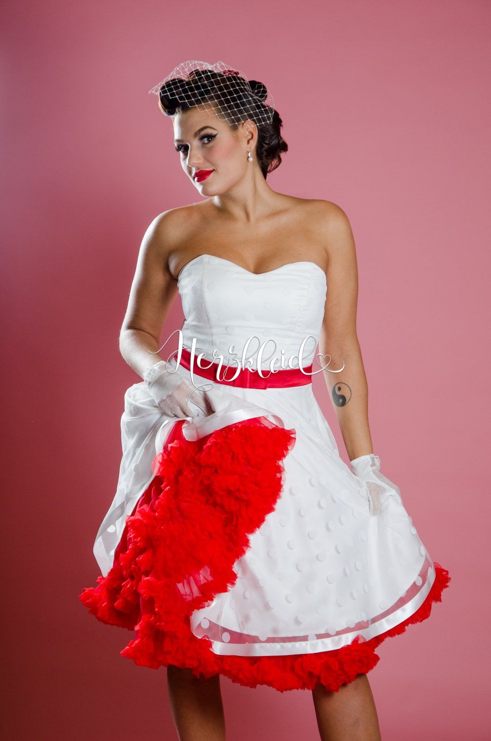 Eine Frau in einem weiß-roten Kleid steht vor einem rosa Hintergrund.