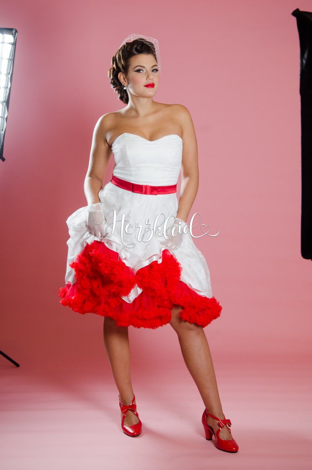 Eine Frau in einem weiß-roten Kleid steht vor einem rosa Hintergrund.