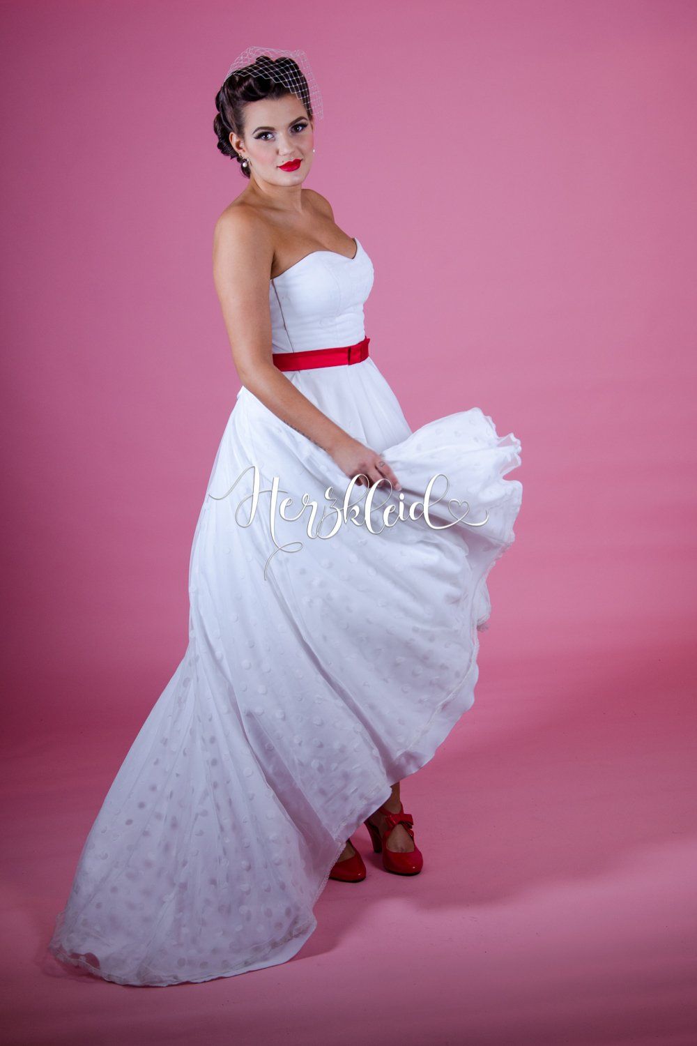 Eine Frau in einem weißen Brautkleid mit einer roten Schärpe steht vor einem rosa Hintergrund.