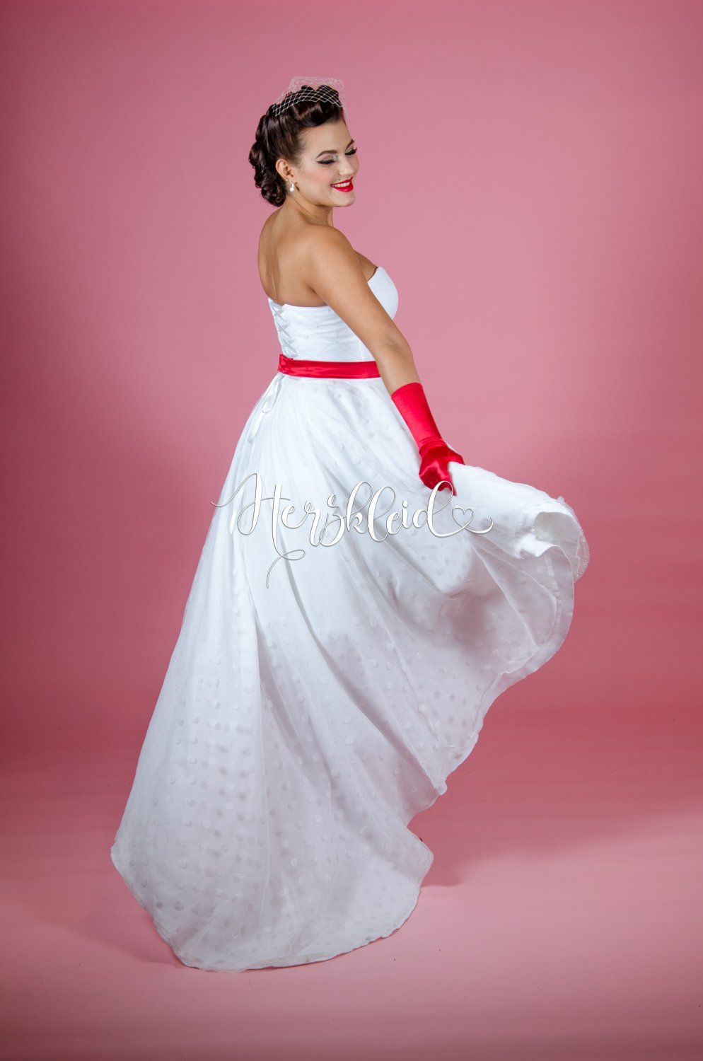 Eine Frau in einem weißen Kleid und roten Handschuhen tanzt vor einem rosa Hintergrund.