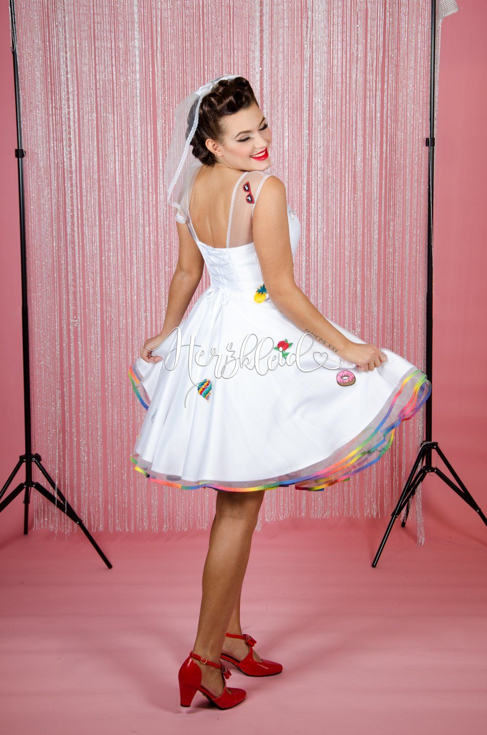 Eine Frau in einem weißen Kleid mit einem Regenbogenrock steht vor einem rosa Vorhang.