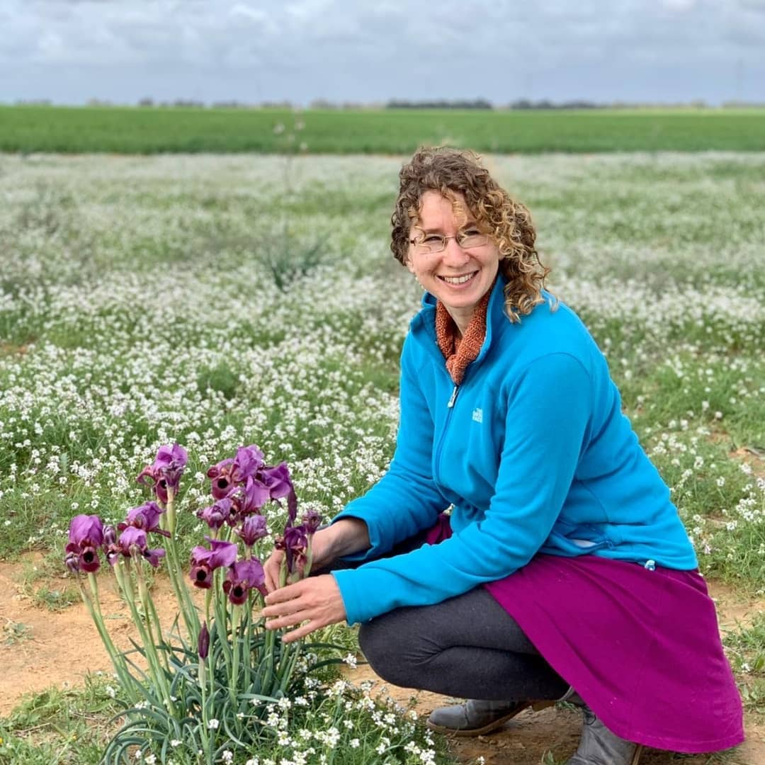 Woman in blue jacket and purple skirt crouching by purple flowers in a field of white flowers.