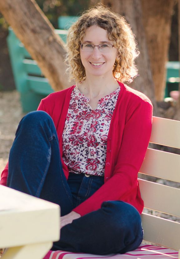 Woman in red sweater and floral shirt smiles, sitting on bench outdoors.