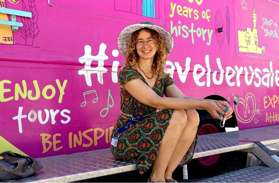 Woman in floral dress and hat smiles, seated in front of a colorful bus with travel-themed graffiti.