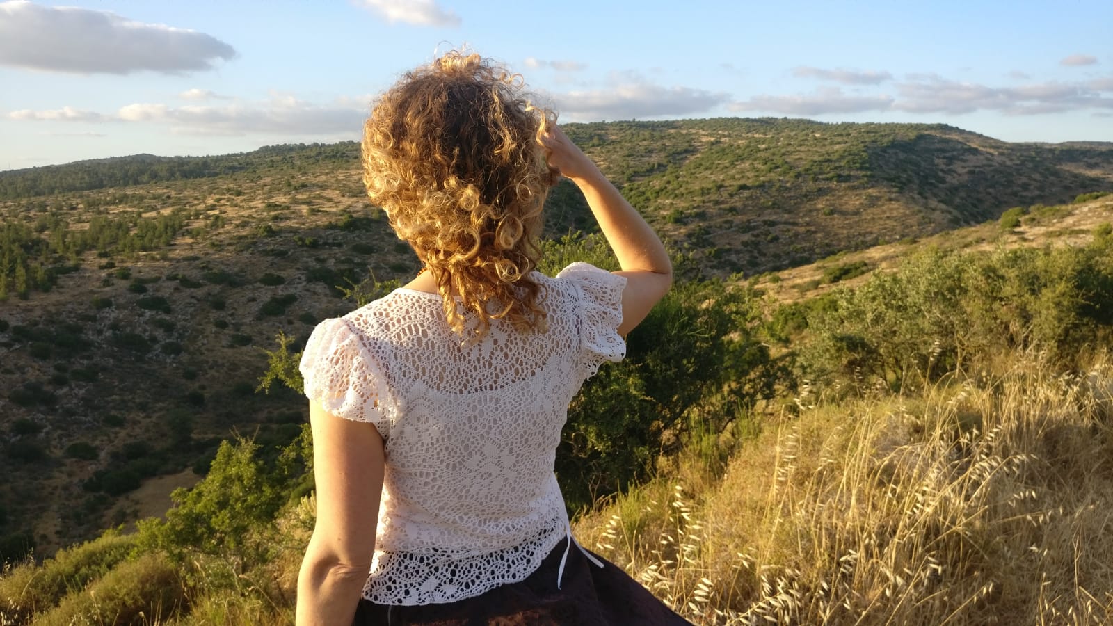 Woman with curly hair looking out at a hilly landscape, arm raised, wearing a white lace top.