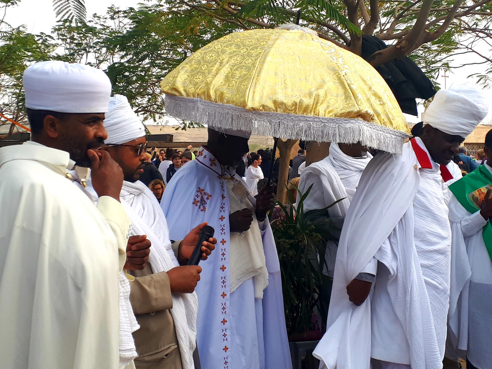 Clergy in white robes, some with head coverings, holding a golden umbrella during an outdoor ceremony.
