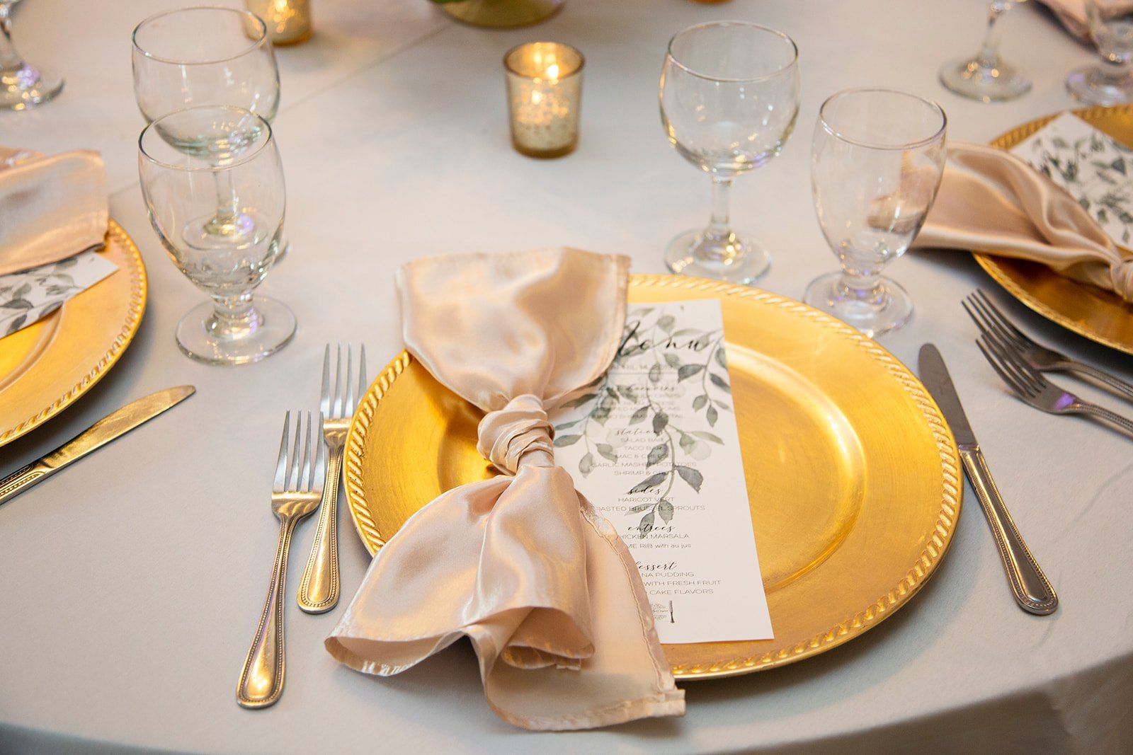 Elegant table setting with gold chargers, beige napkins, glassware, and candlelight on a white tablecloth
