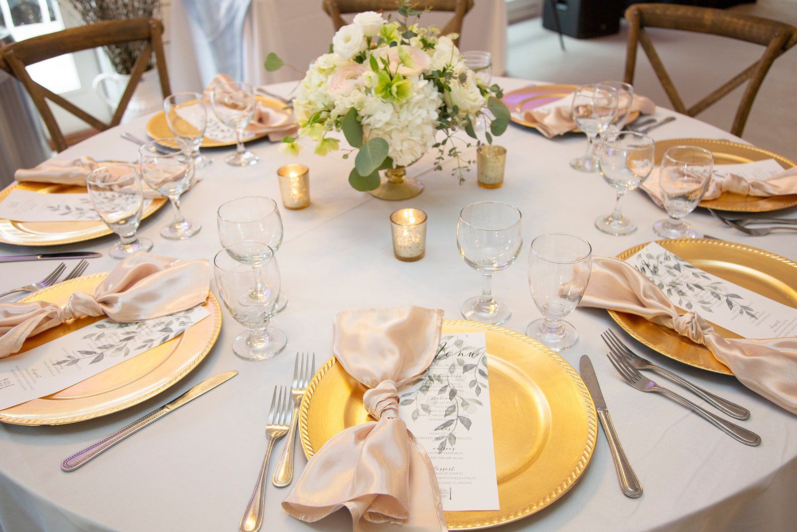 Elegant round dining table set with gold accents, white flowers, candles, and folded napkins on cream plates