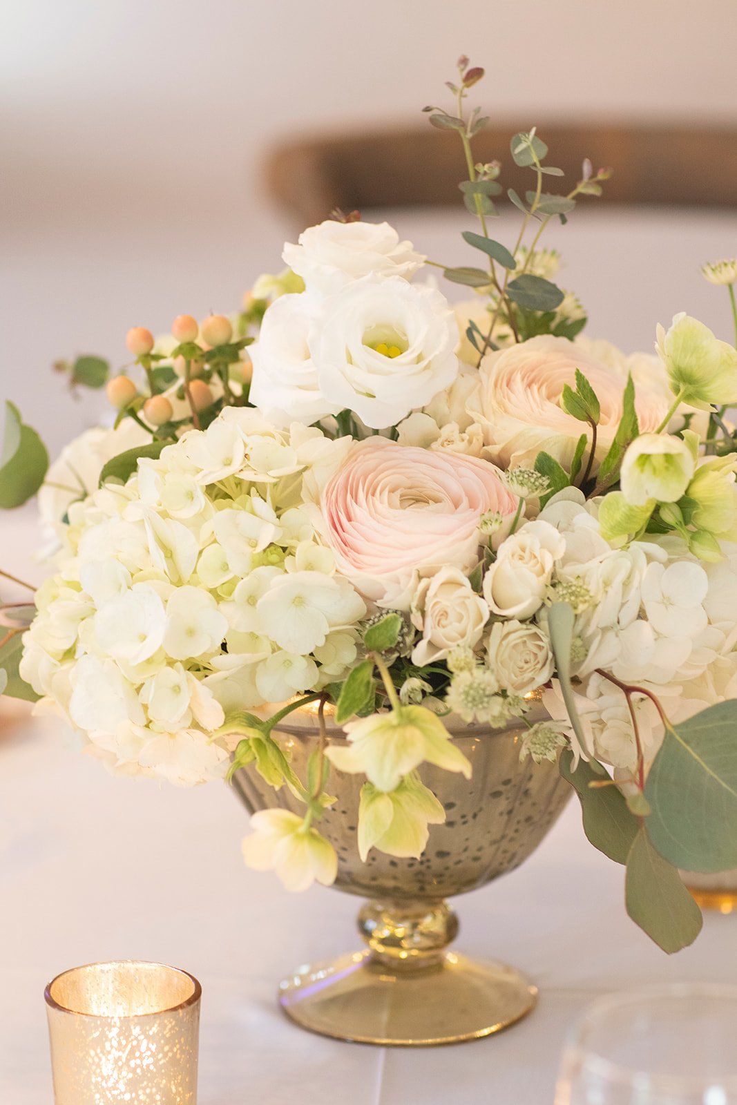White and blush floral arrangement in a gold vase beside a lit candle on a soft background
