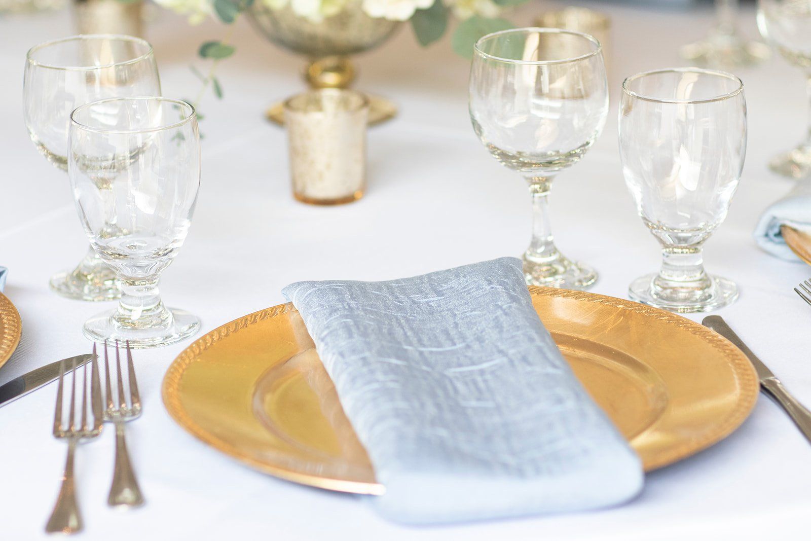 Elegant dining table set with gold chargers, crystal glasses, silverware, and a white napkin