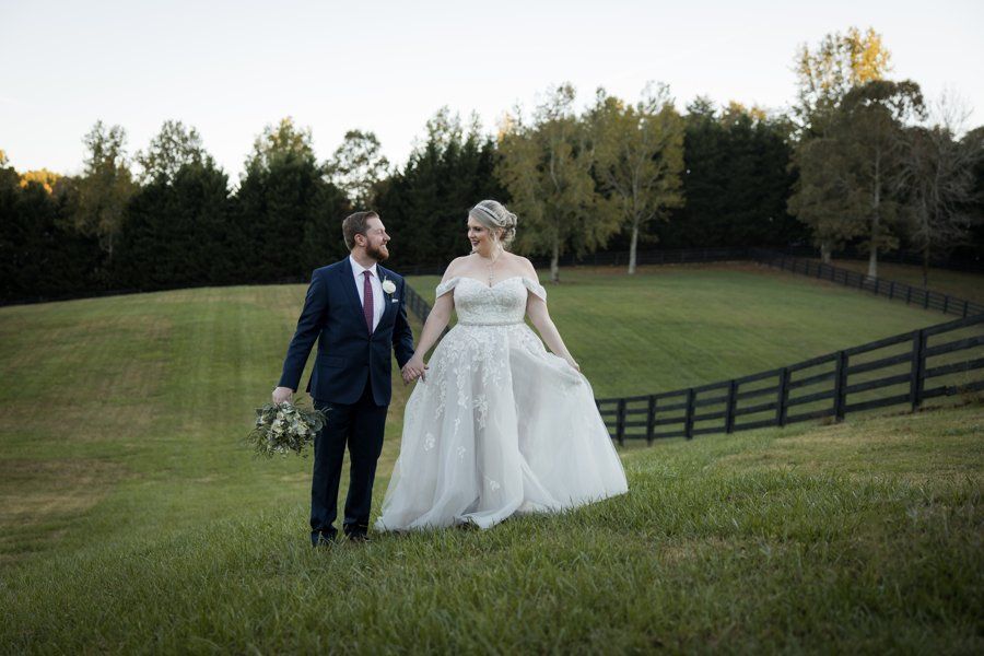 A bride and groom are walking through a grassy field holding hands.
