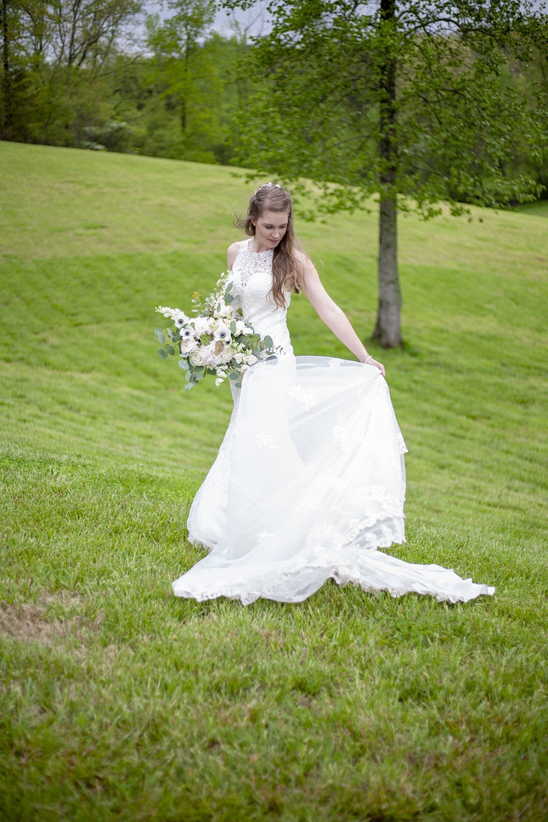 Bride in white gown holding bouquet on a grassy hillside under trees