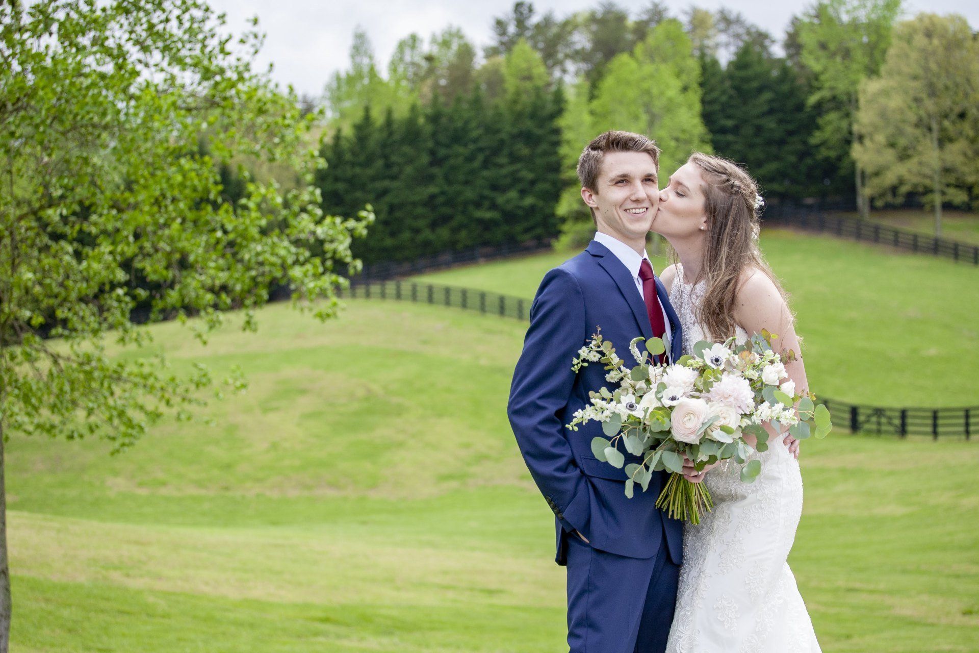 Bride and groom standing on a green lawn, holding a white bouquet, with trees in the background