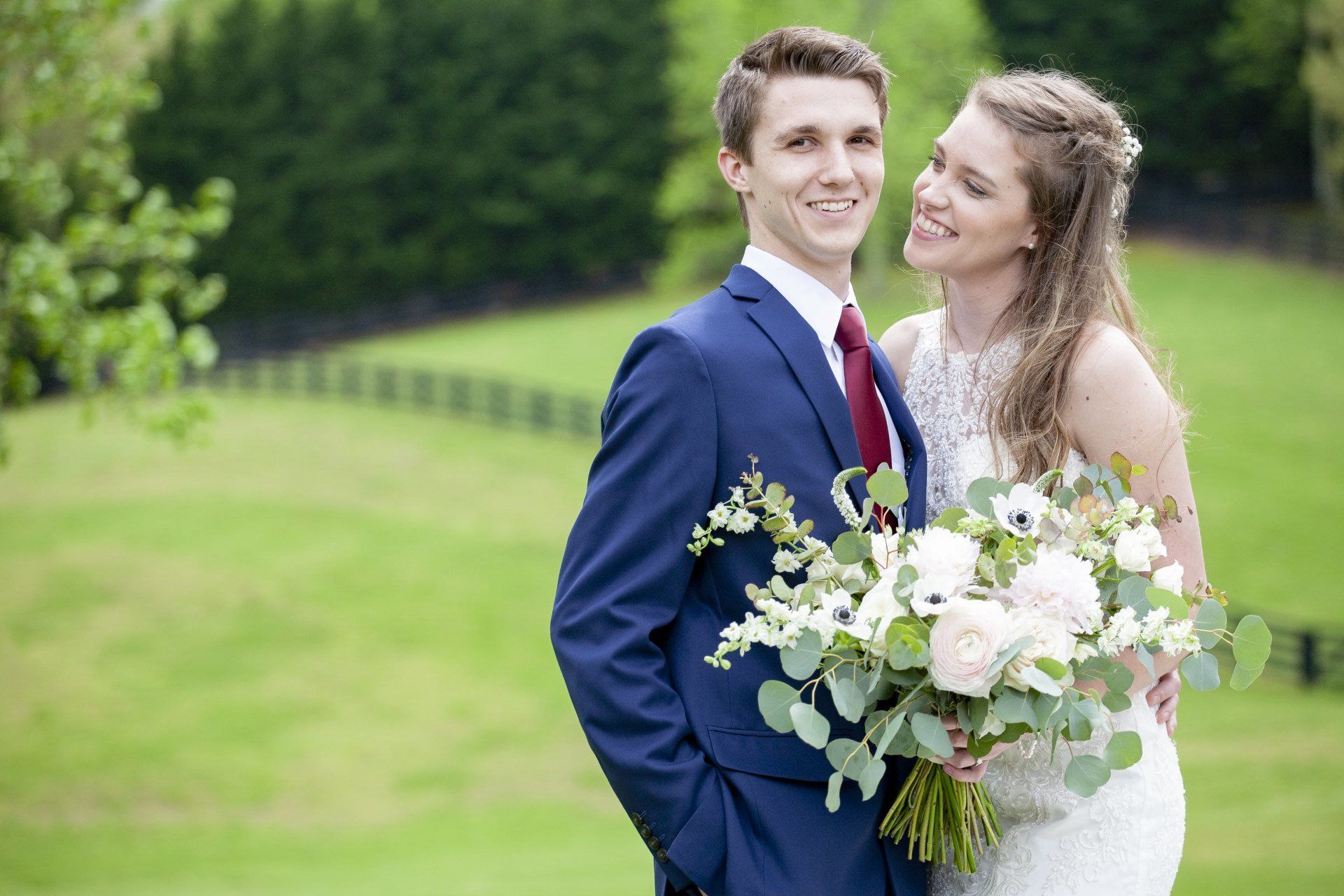 Bride and groom smiling outdoors, holding a bouquet in a green garden.