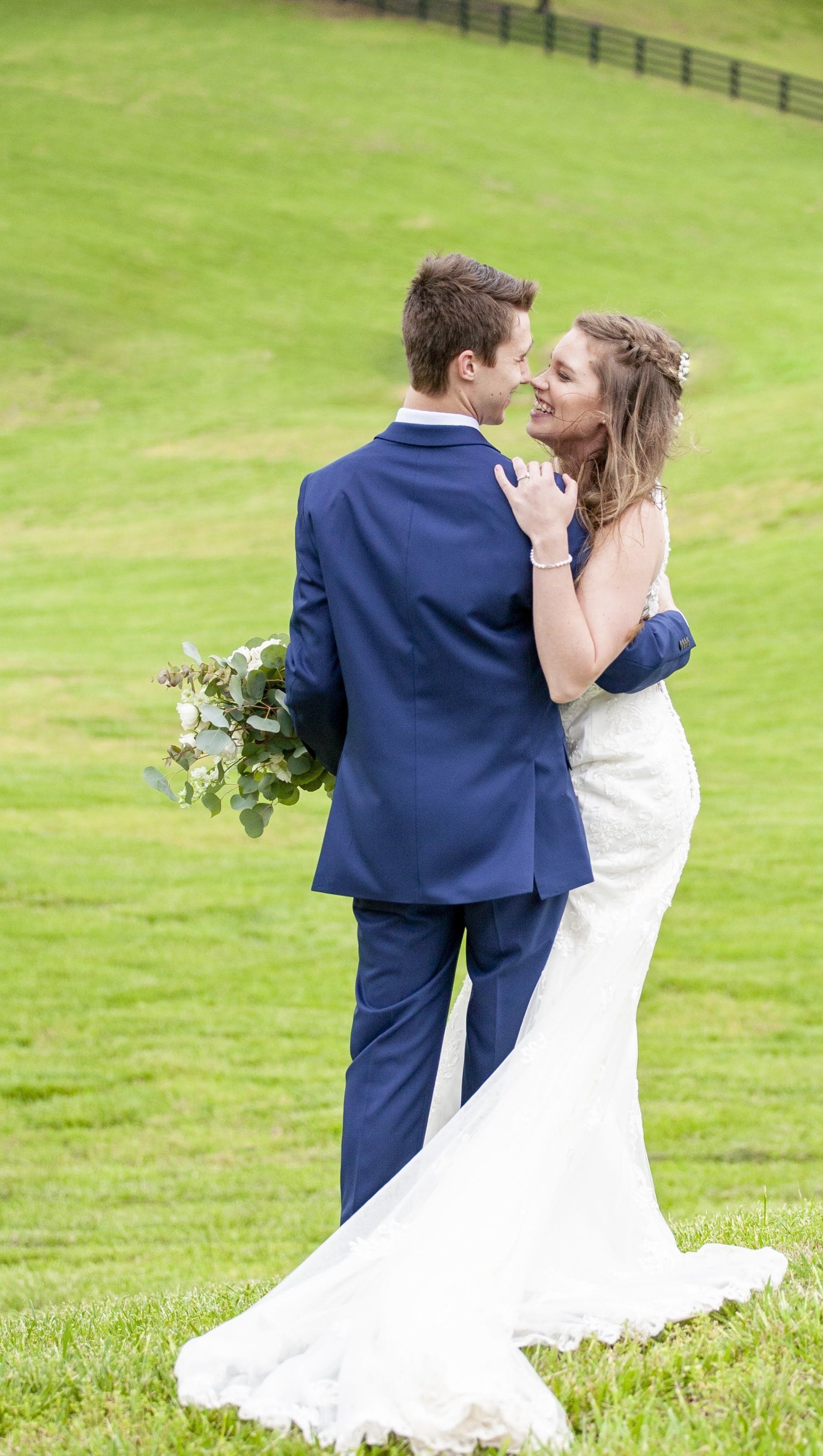 Couple embracing on a grassy hillside, with the woman in a white wedding dress and the man in a blue suit.
