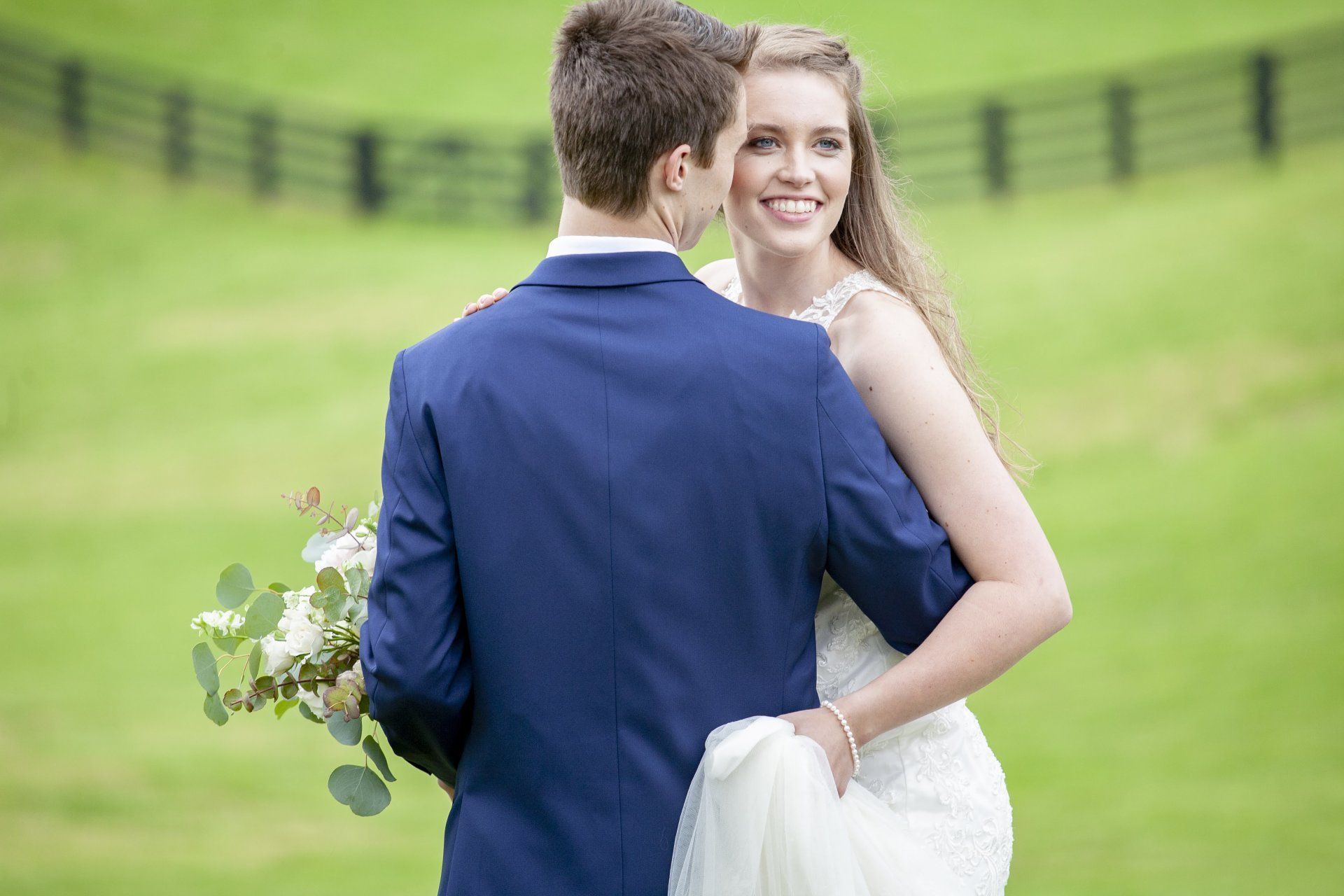 Couple embracing outdoors on a grassy field, with the man in a navy suit holding a bouquet.