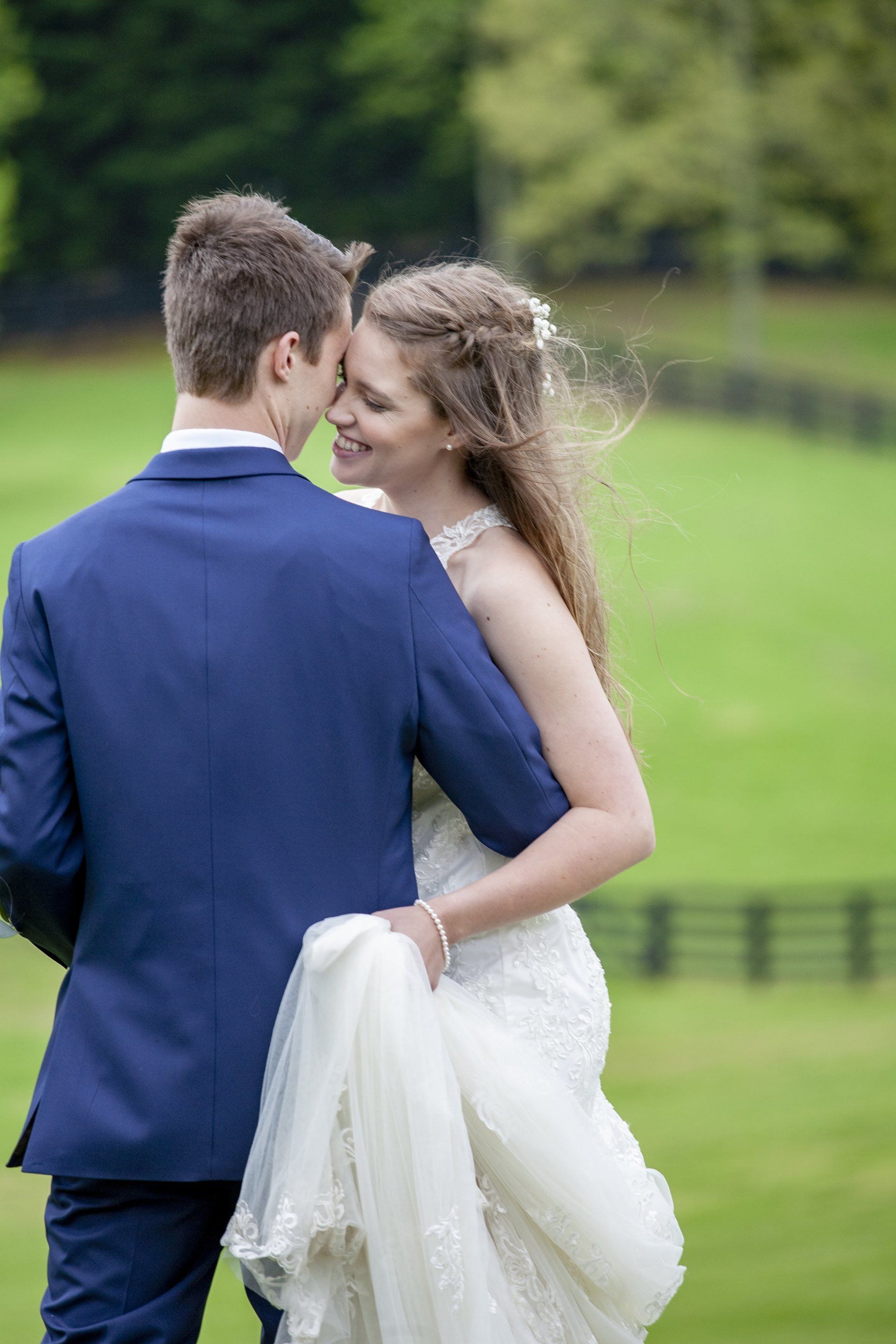 Bride and groom embrace outdoors on a green lawn, with the bride in a white dress and veil and the groom in a blue suit