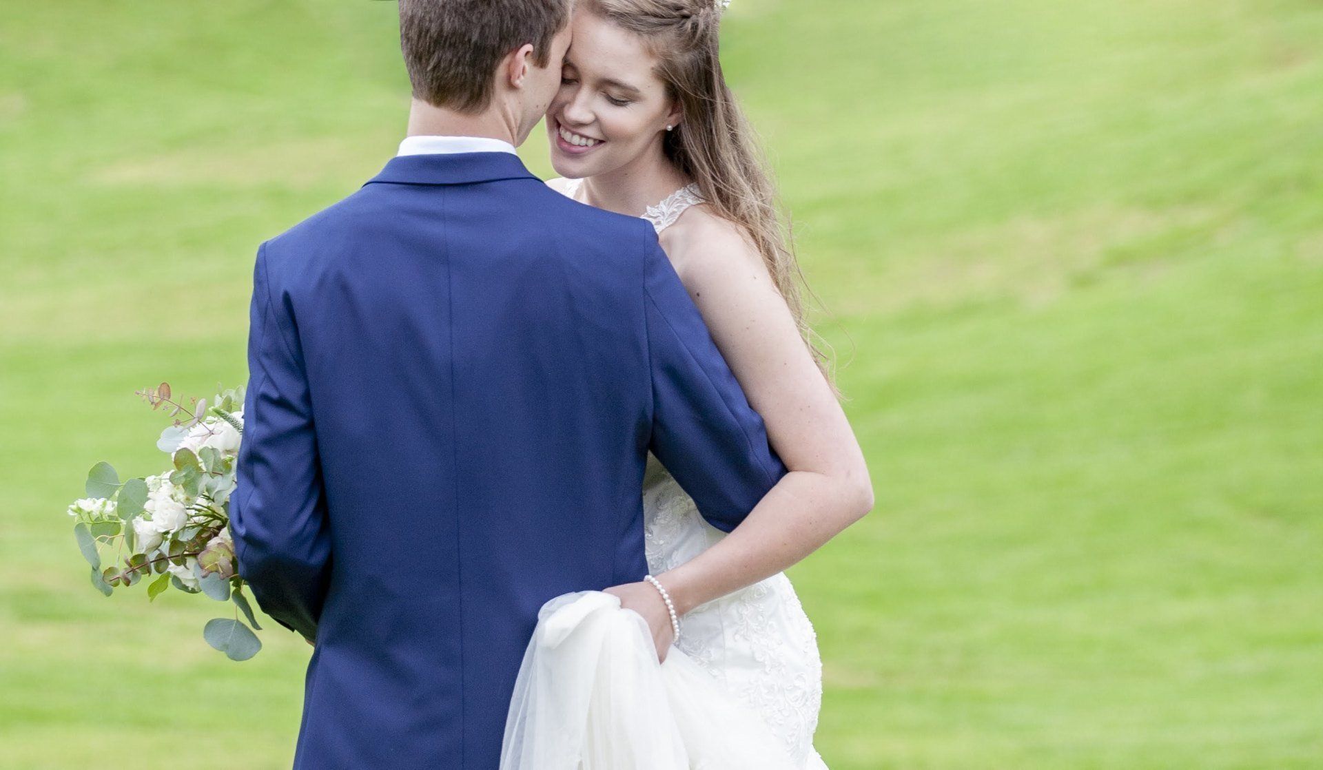 Bride and groom embracing outdoors on a grassy lawn, the bride smiling and holding a bouquet