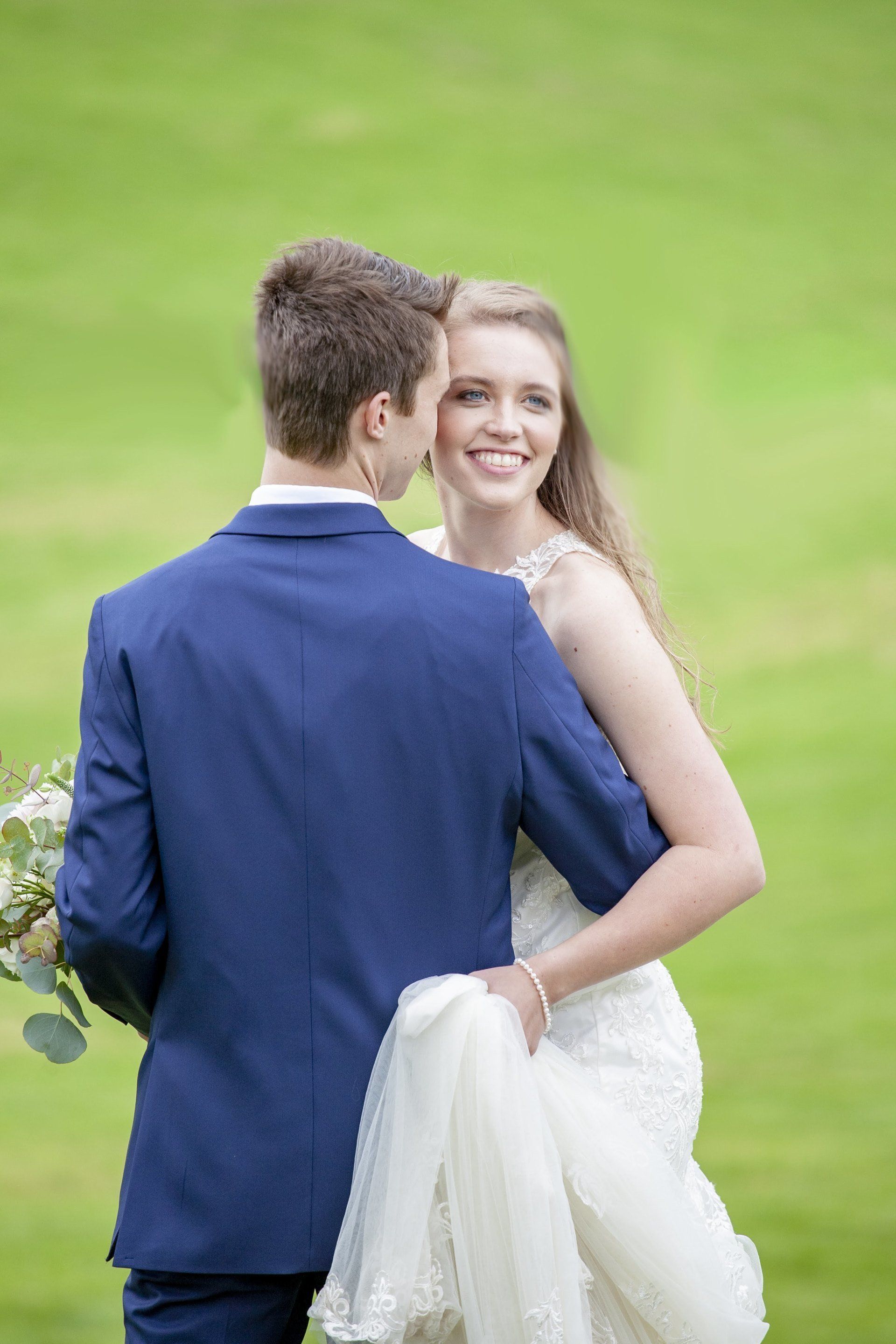 Bride and groom posing outdoors, smiling in a green field, with the bride holding her white dress.