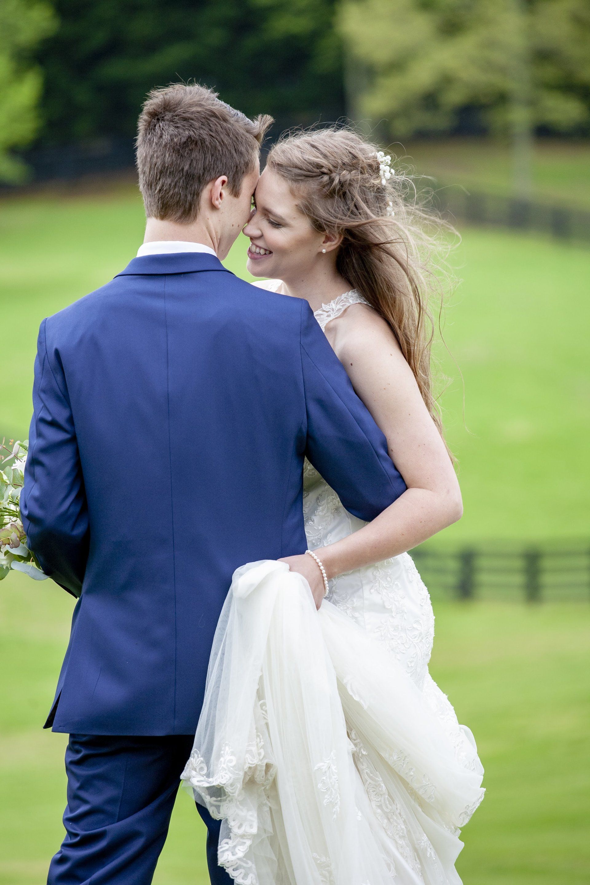 Couple in formal attire embracing outdoors in a green park, with the woman holding a white dress train