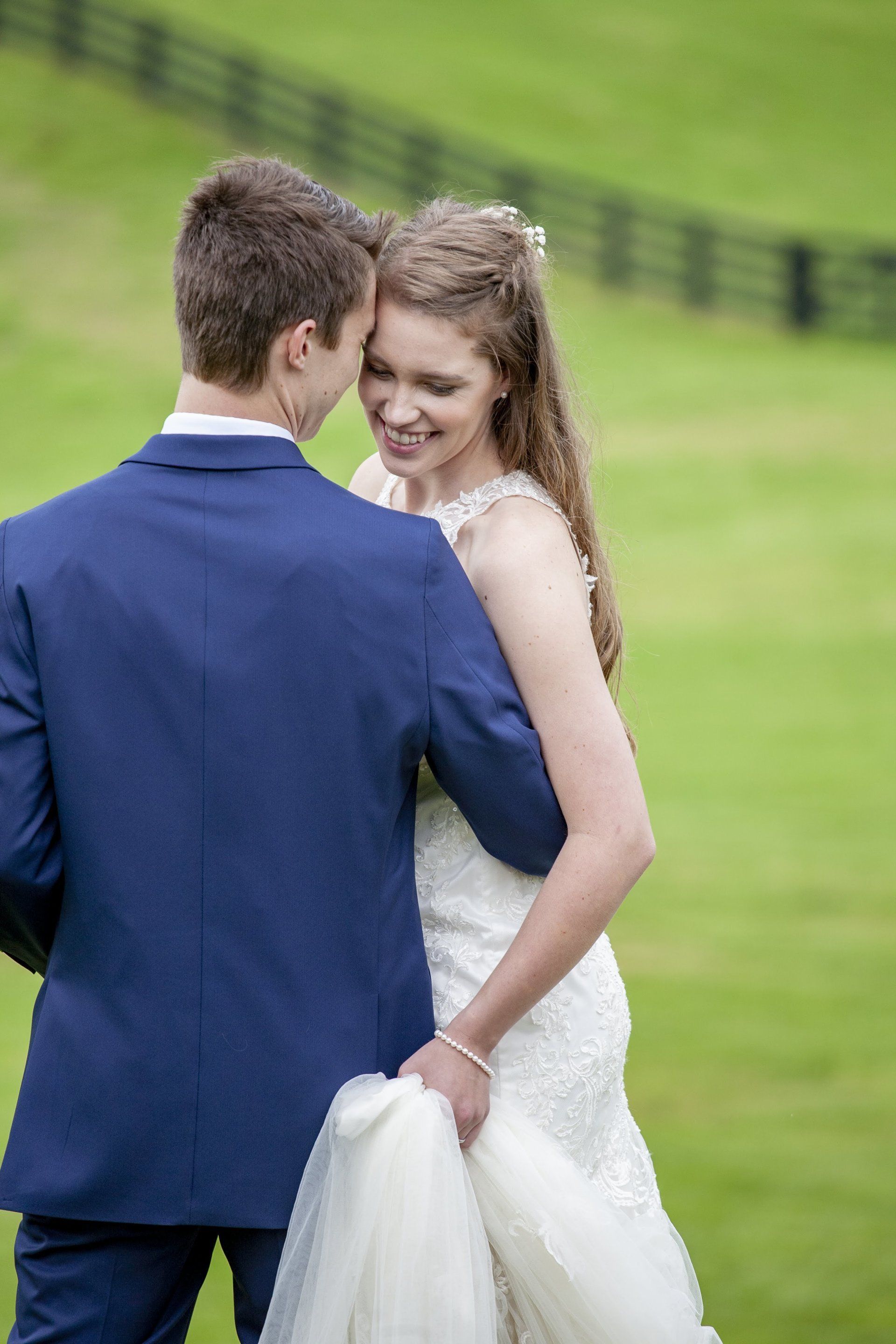 Bride and groom embracing outdoors on a green hillside, the bride in a white gown and the groom in a blue suit