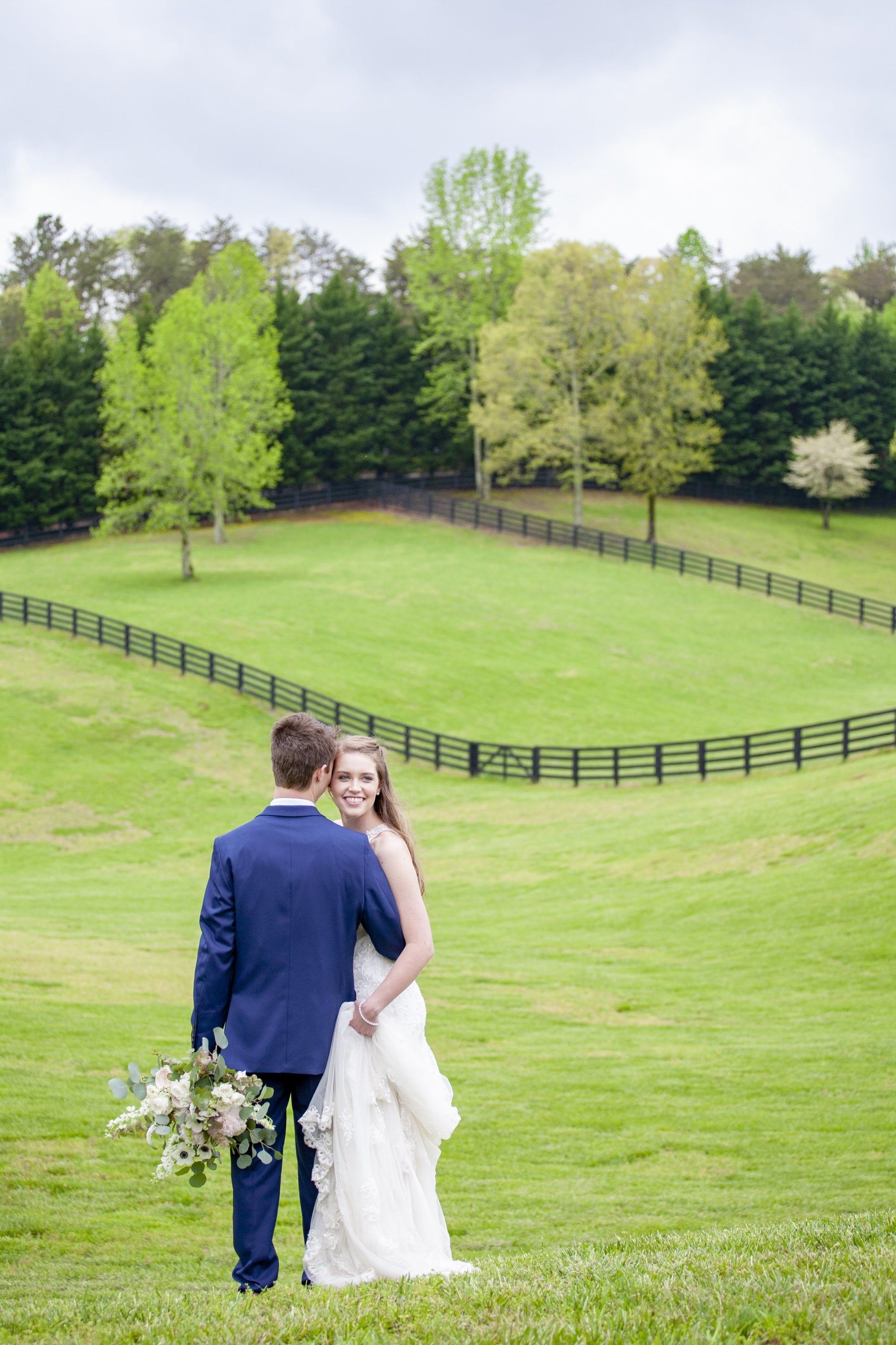Couple in wedding attire standing on a green hillside with a dark wooden fence and trees behind them