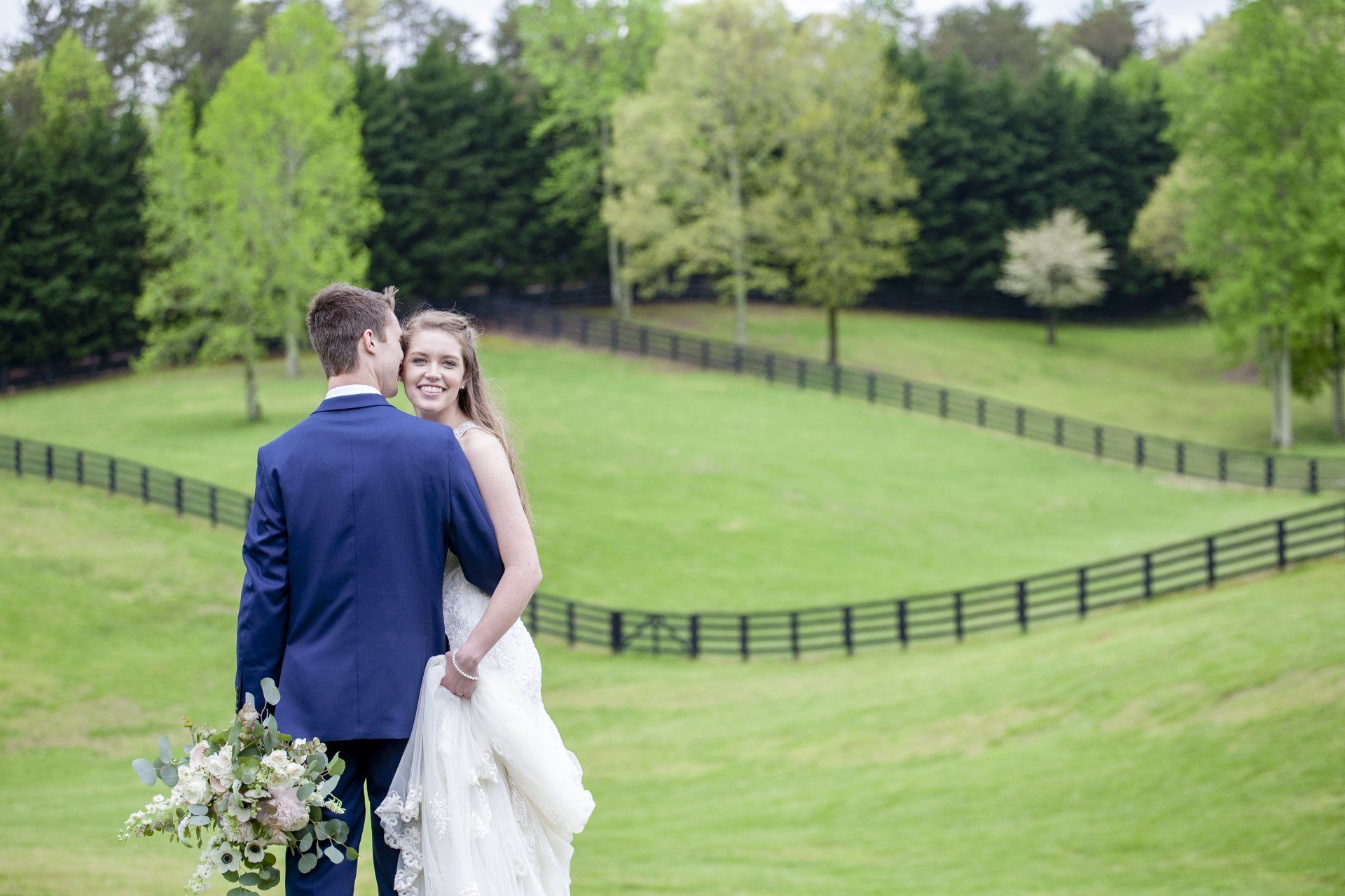 Couple in wedding attire standing in a green field with trees and a fence in the background