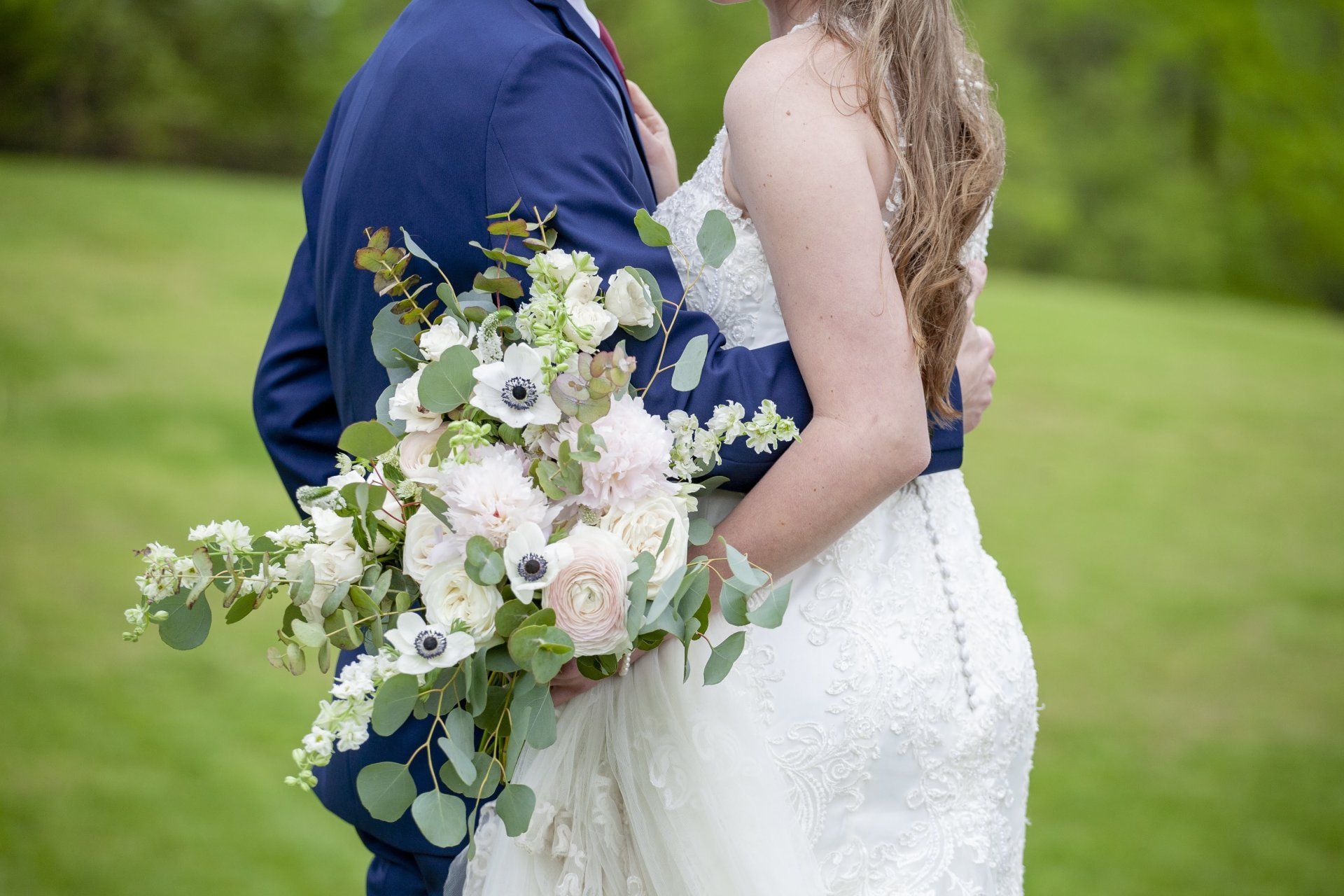 Bride and groom embracing in a grassy field, holding a white and green bouquet.