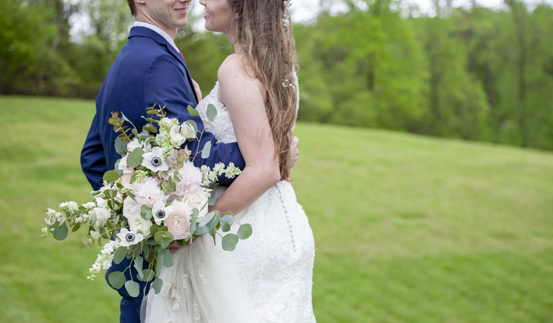 Bride and groom embracing outdoors, holding a white and purple bouquet on a green lawn