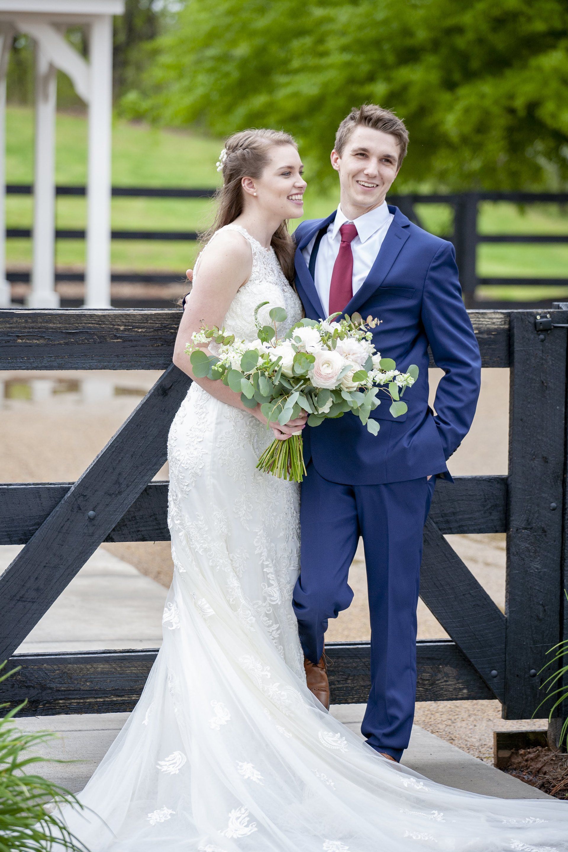 Wedding couple posing outdoors by a black fence, bride in white gown holding bouquet, groom in blue suit
