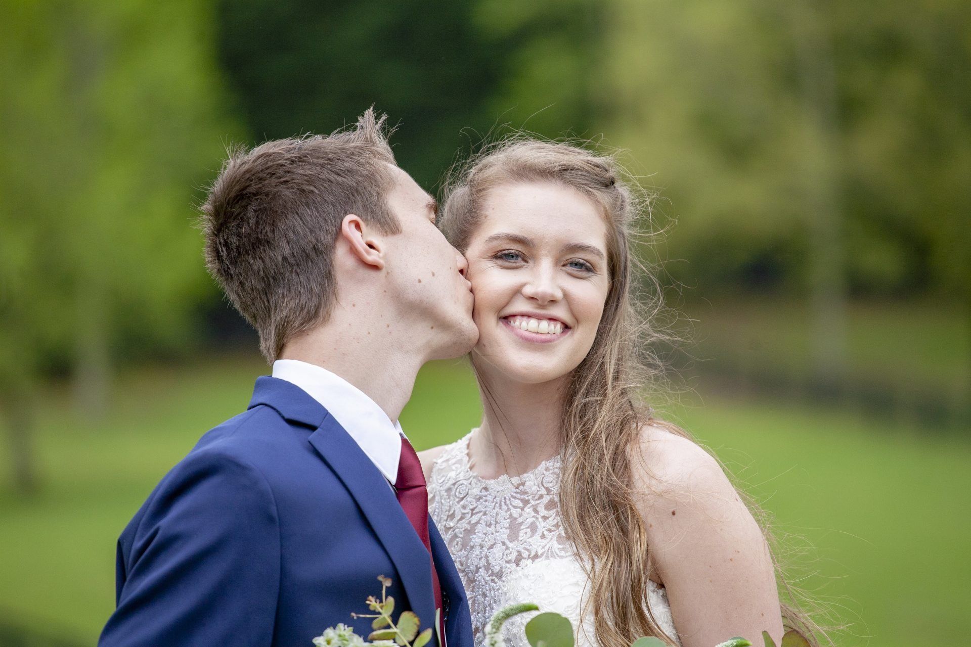 Man in a blue suit kissing a smiling woman in a white dress outdoors