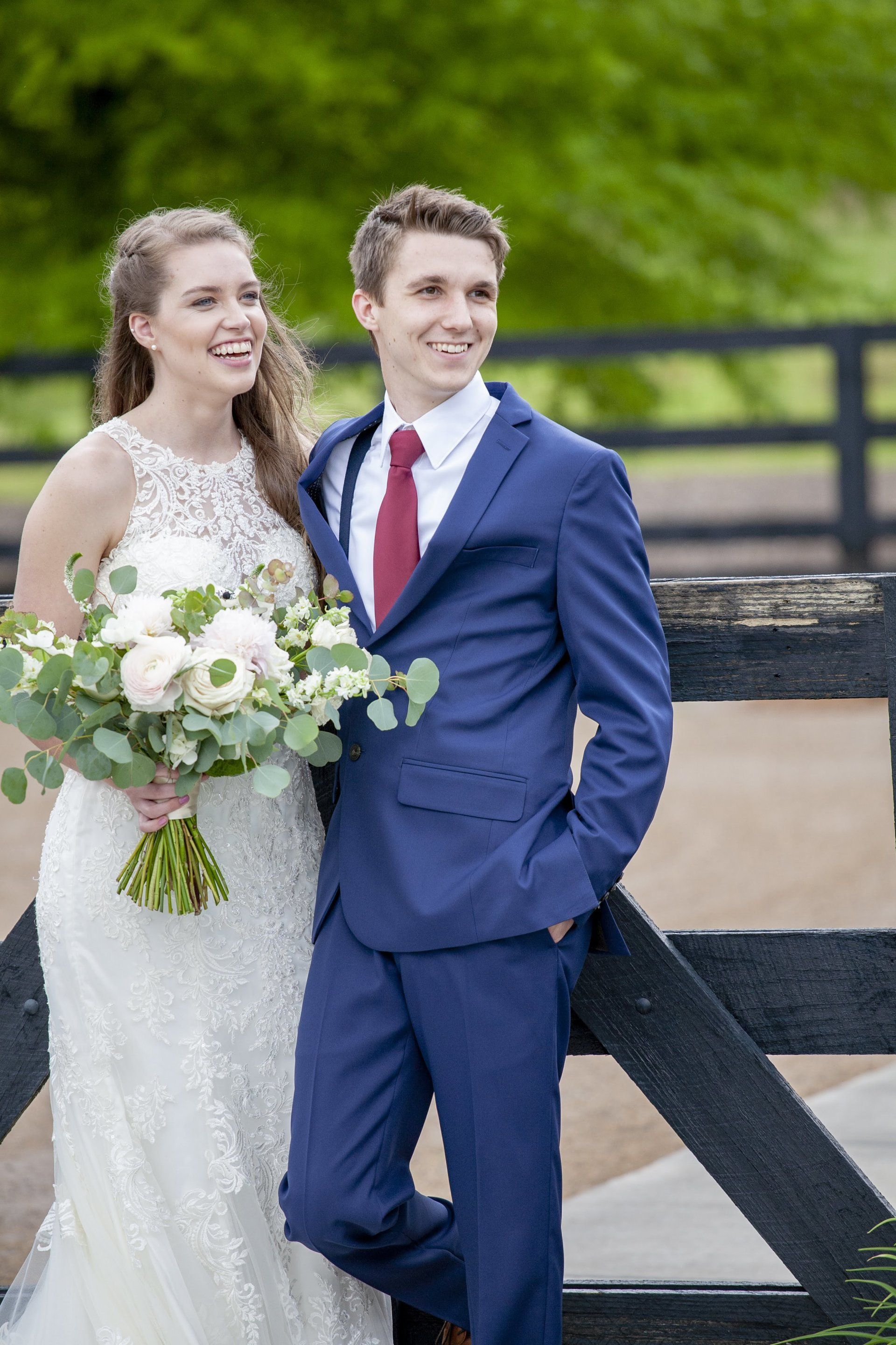 Couple in wedding attire standing by a wooden fence, smiling outdoors with green trees in the background.