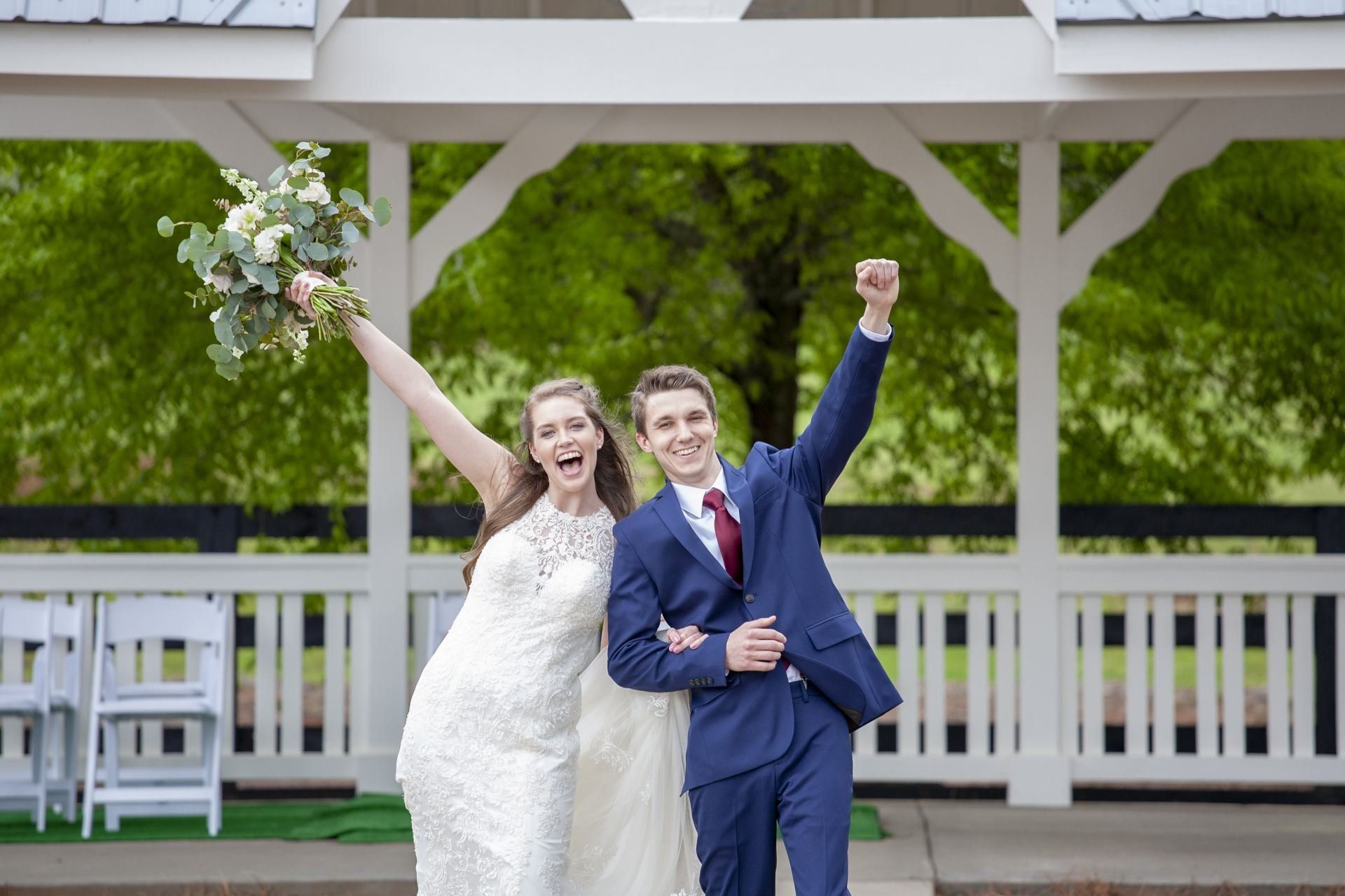 Smiling bride and groom in white and blue outfits, celebrating with raised arms in a gazebo.