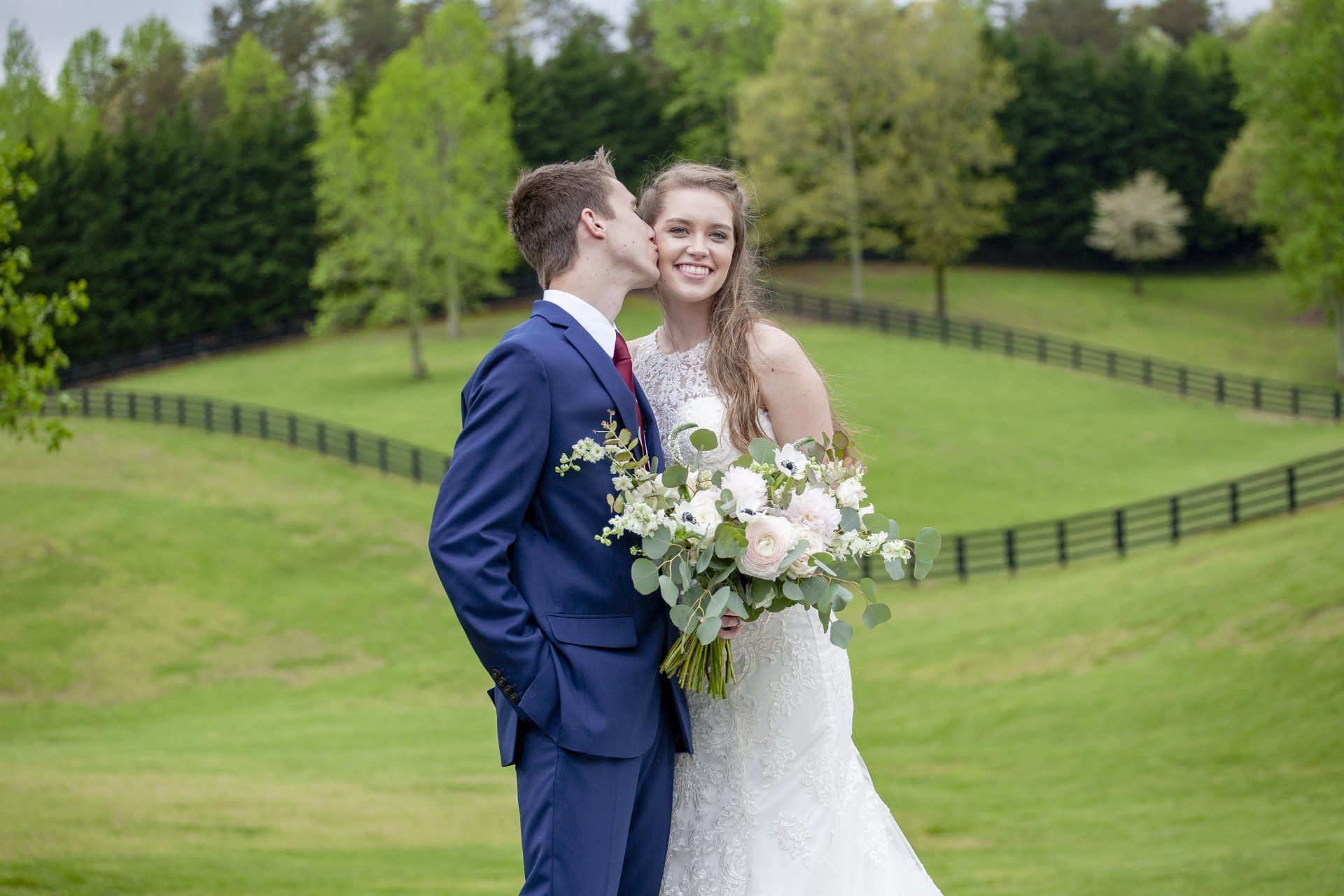Bride and groom embrace in a green field, the bride holding a white bouquet.