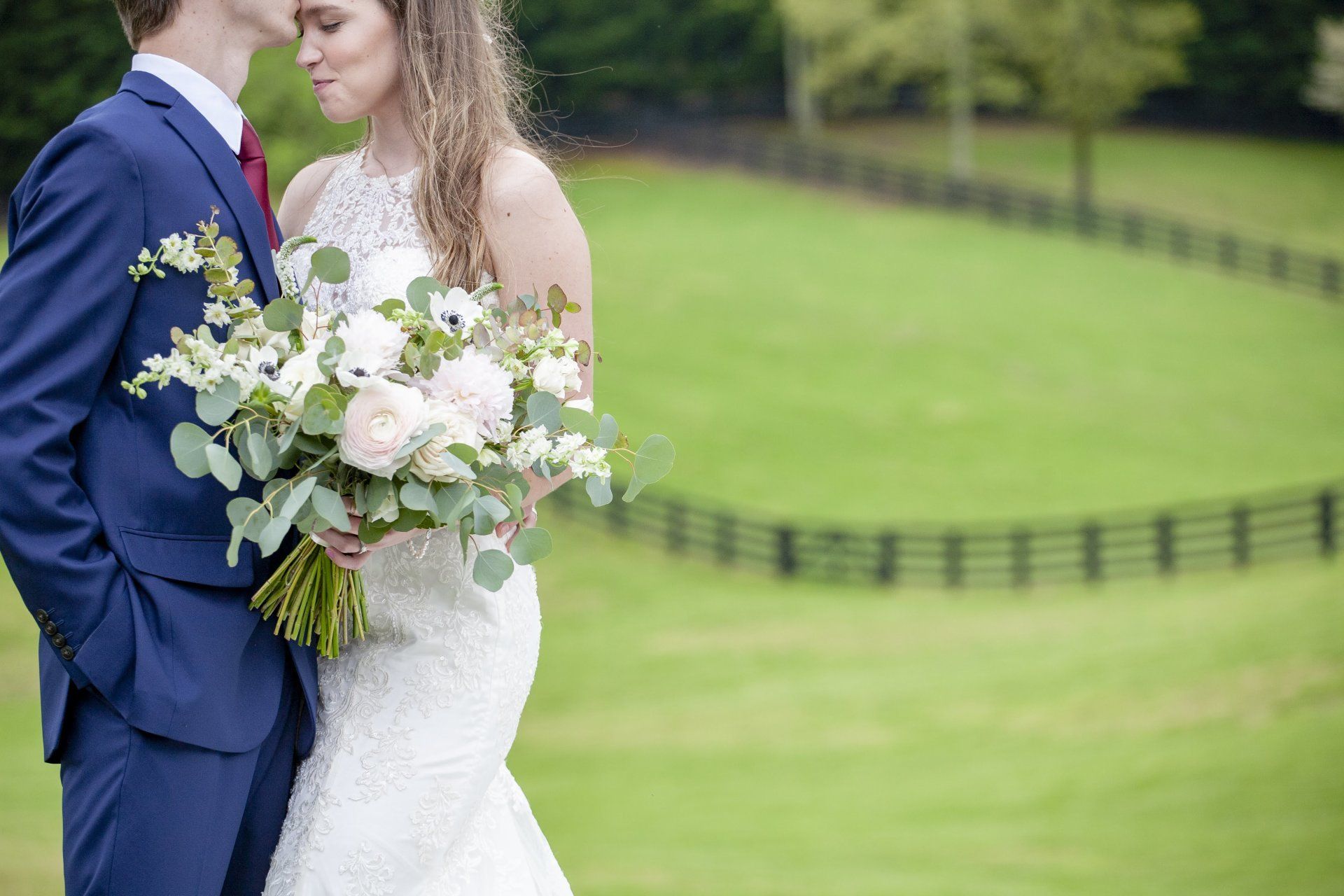 Bride and groom embracing outdoors, bride holding a white bouquet on a green lawn.