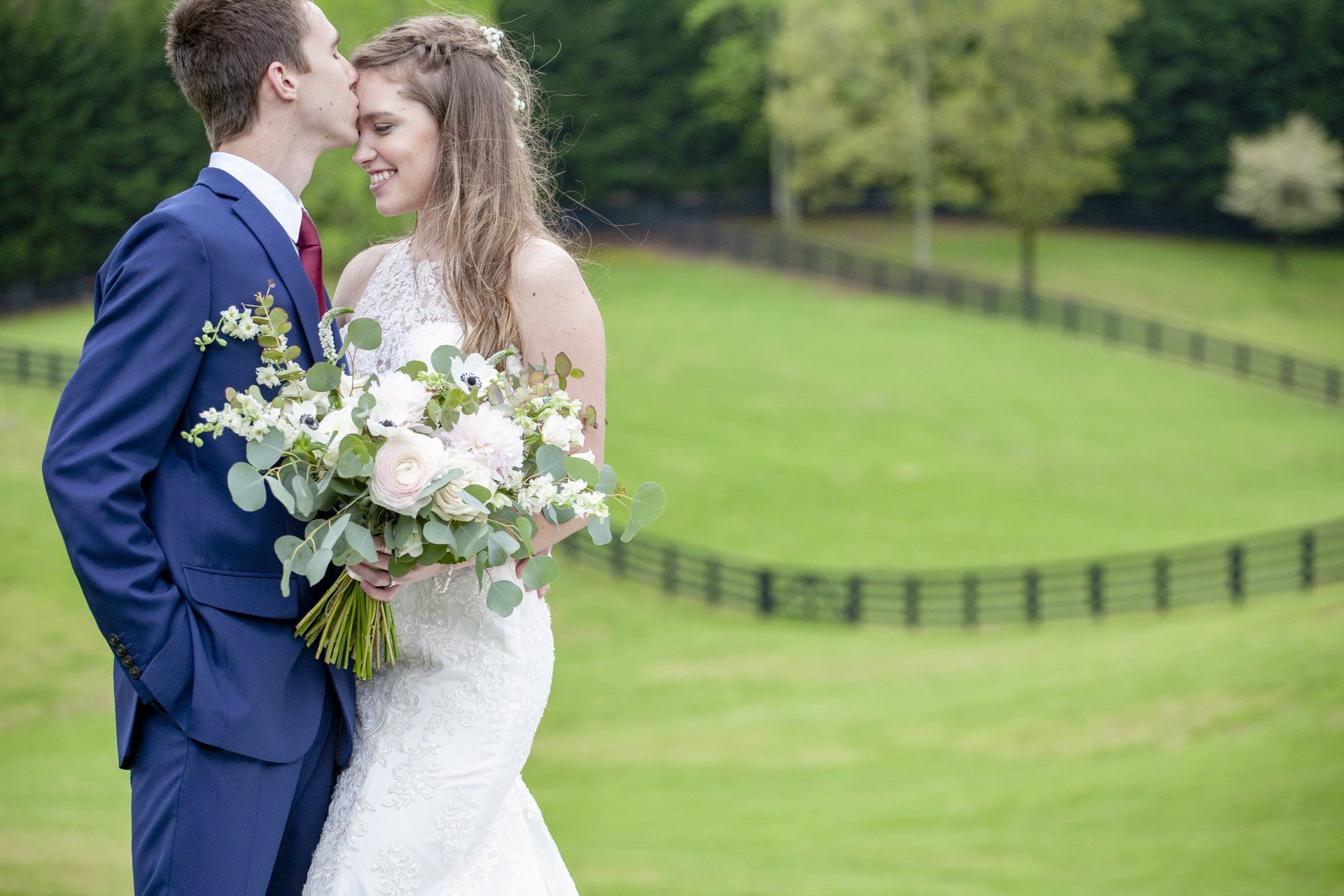 Newlywed couple embracing in a green outdoor field, bride holding a white bouquet
