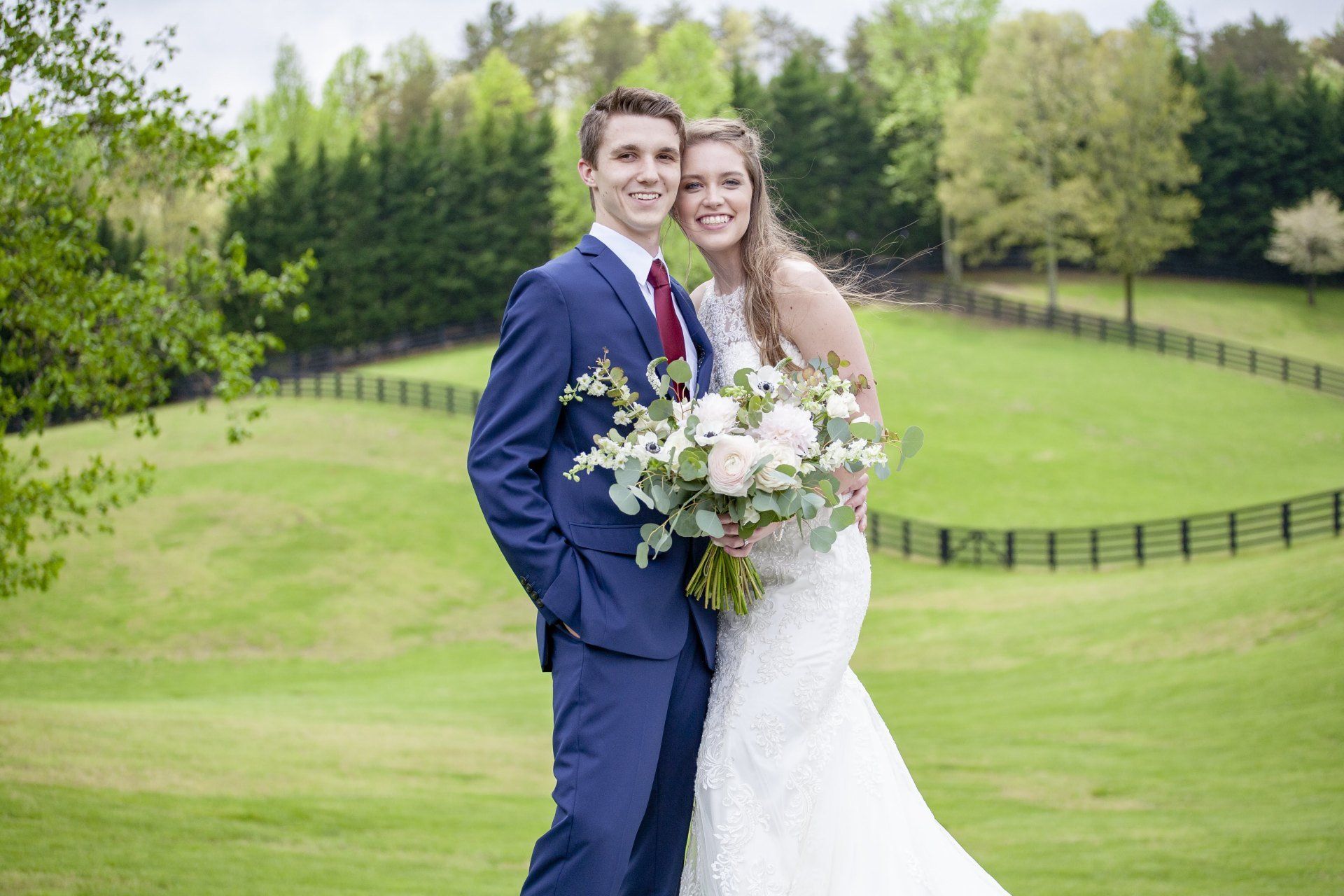 Wedding couple posing on a green hillside with trees and a split-rail fence behind them.