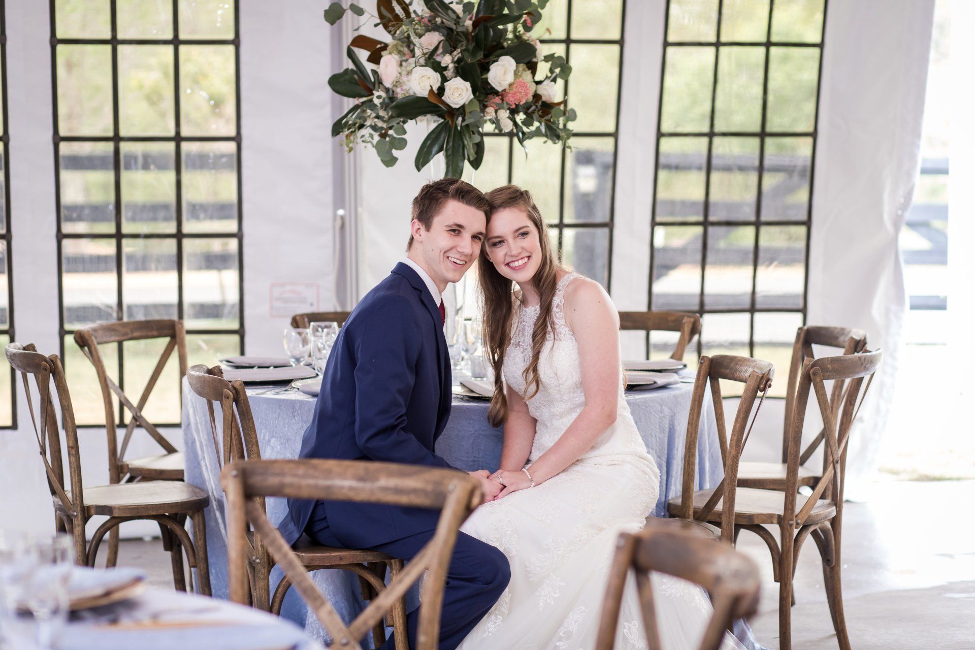 Couple seated at a decorated wedding table, smiling in a bright room with large windows and floral centerpieces