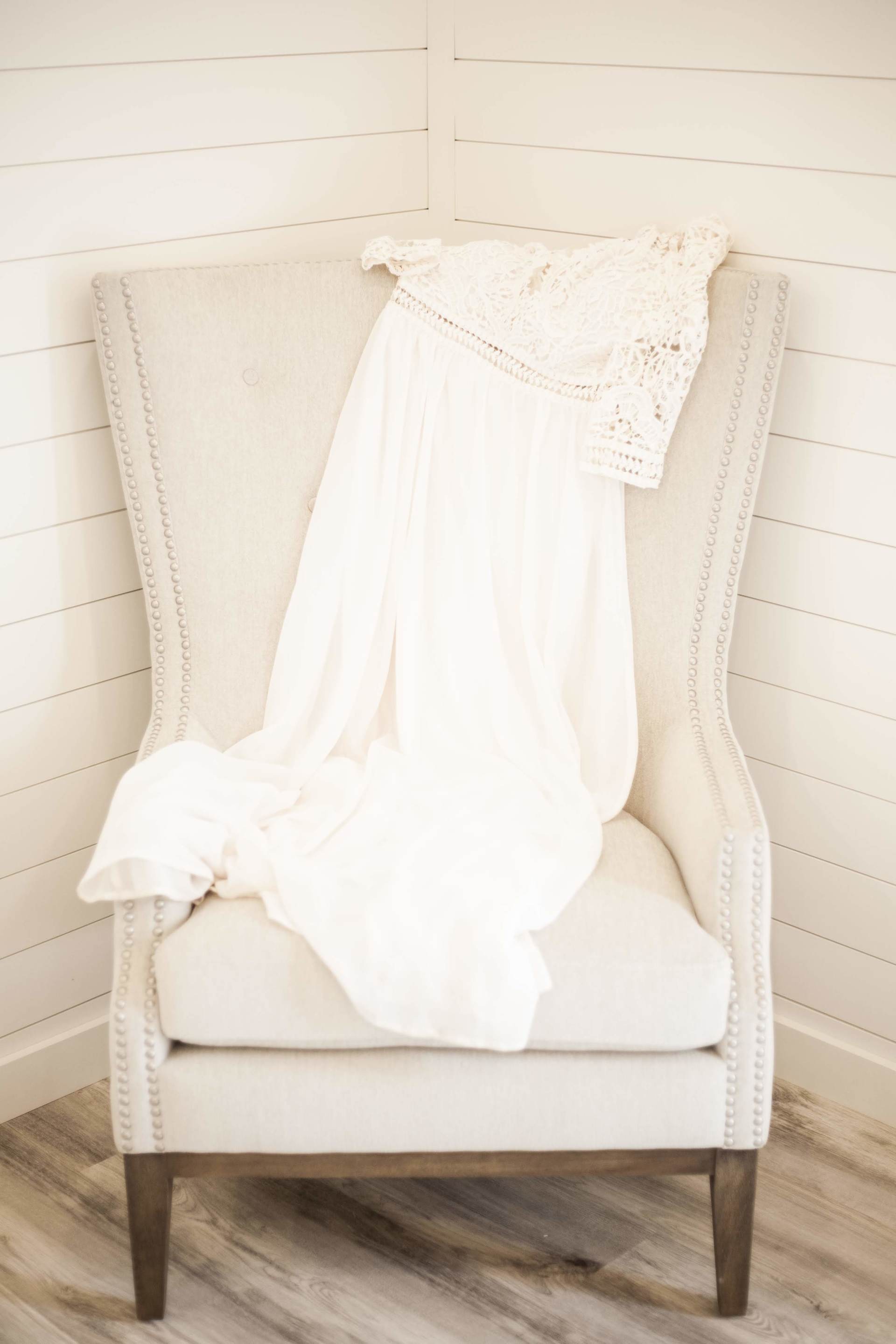 White dress draped over a cream upholstered chair in a bright room with shiplap walls.