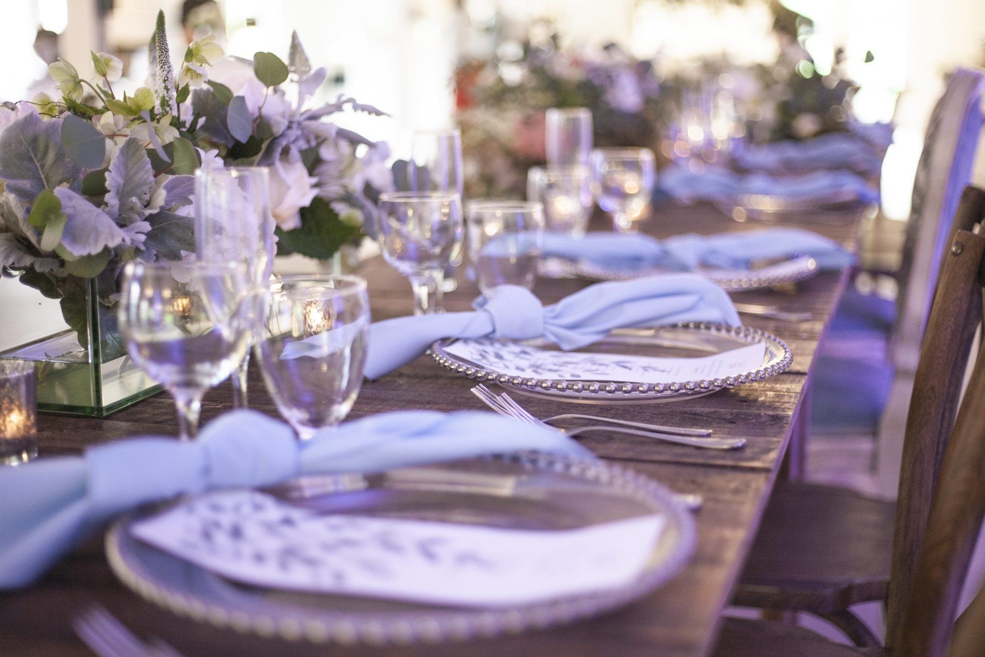 Outdoor dining table set with white plates, folded napkins, glassware, and purple-and-white floral centerpieces