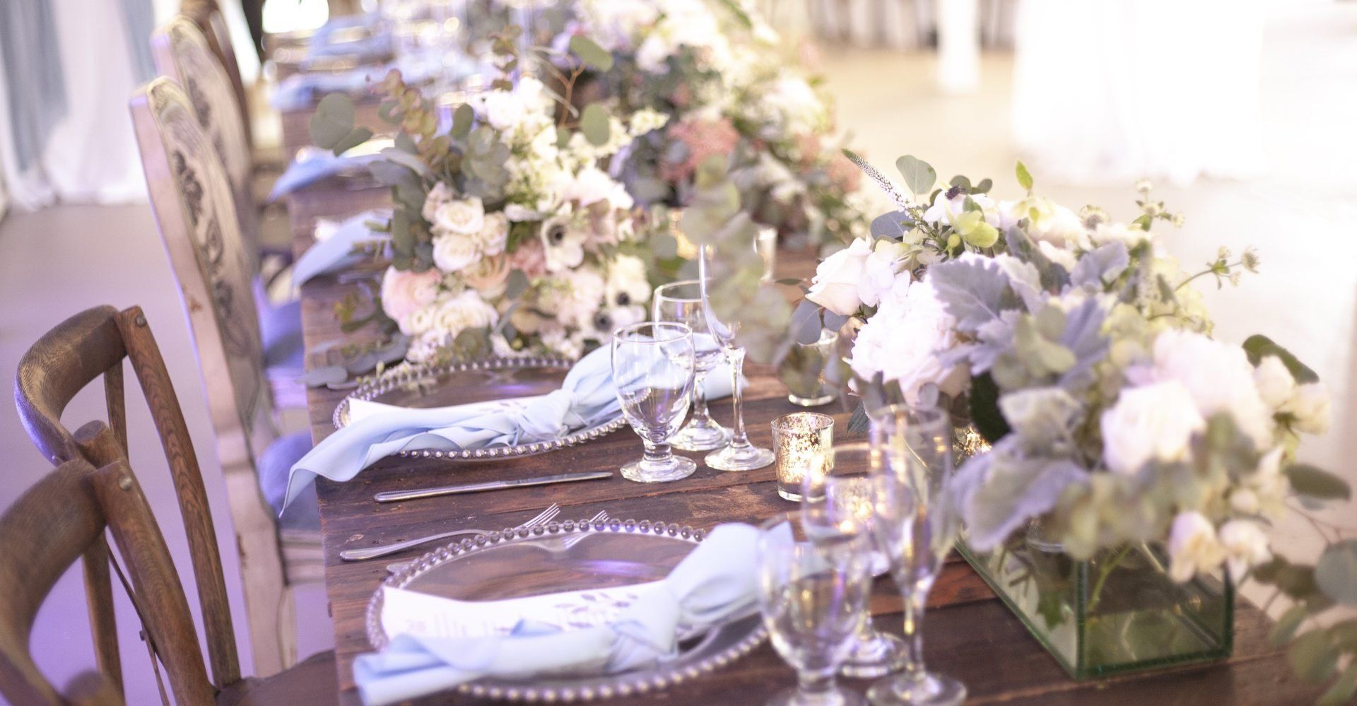 Elegant dining table set with white floral centerpieces, candles, and silver place settings under soft purple lighting