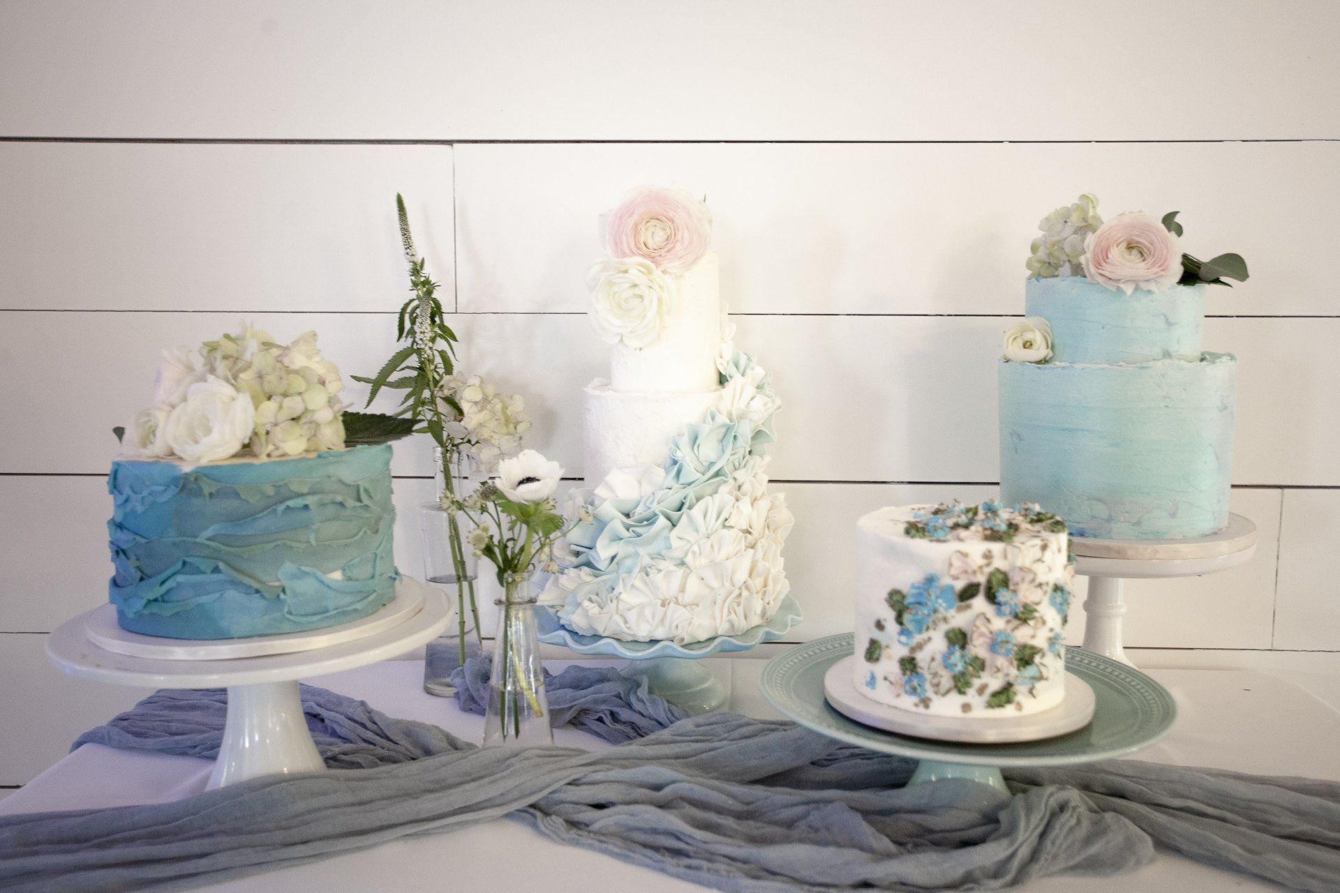 Assorted blue-and-white decorated cakes on stands with white flowers against a white wooden backdrop