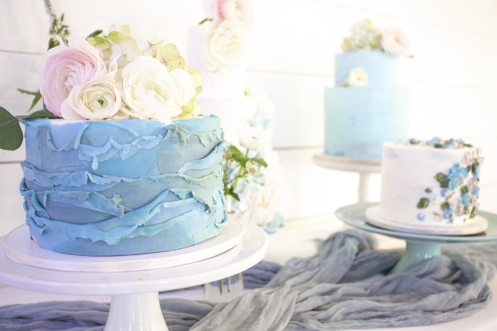 Blue ruffled cake with pastel flowers on a white stand, beside a small decorated cake on a gray drape backdrop